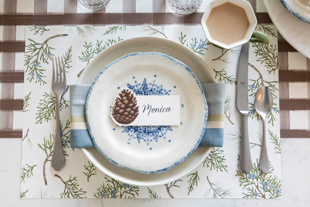 Elegant table setting with the Juniper Sprigs Placemat, a napkin in between two plates and a Pinecone Place Card with the name "Monica" written on it, on top.