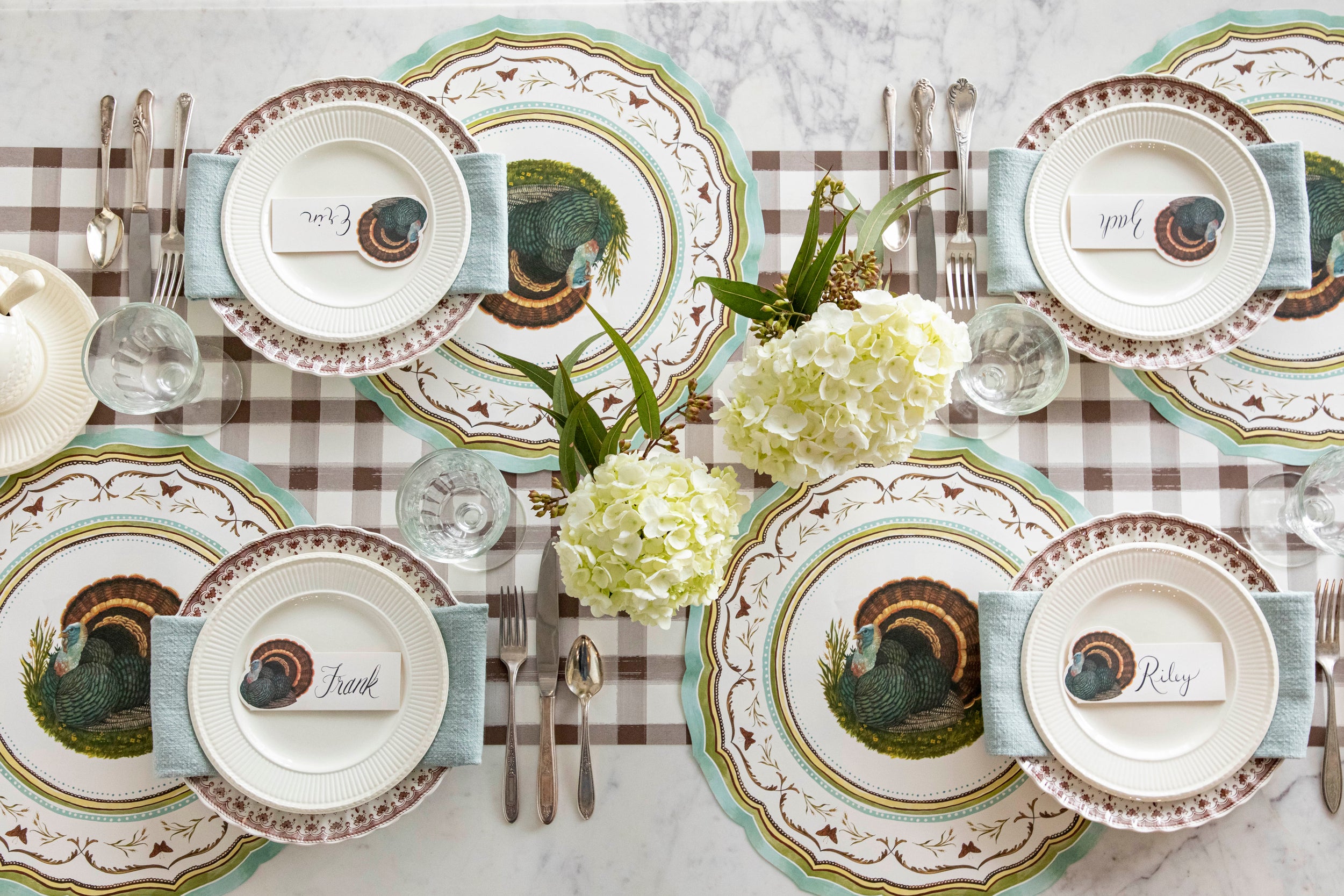 An elegant table setting featuring the Die-cut Heritage Turkey China Placemats, Heritage Turkey Place Cards atop plates, vintage dinnerware and flowers on the Brown Painted Check Runner.