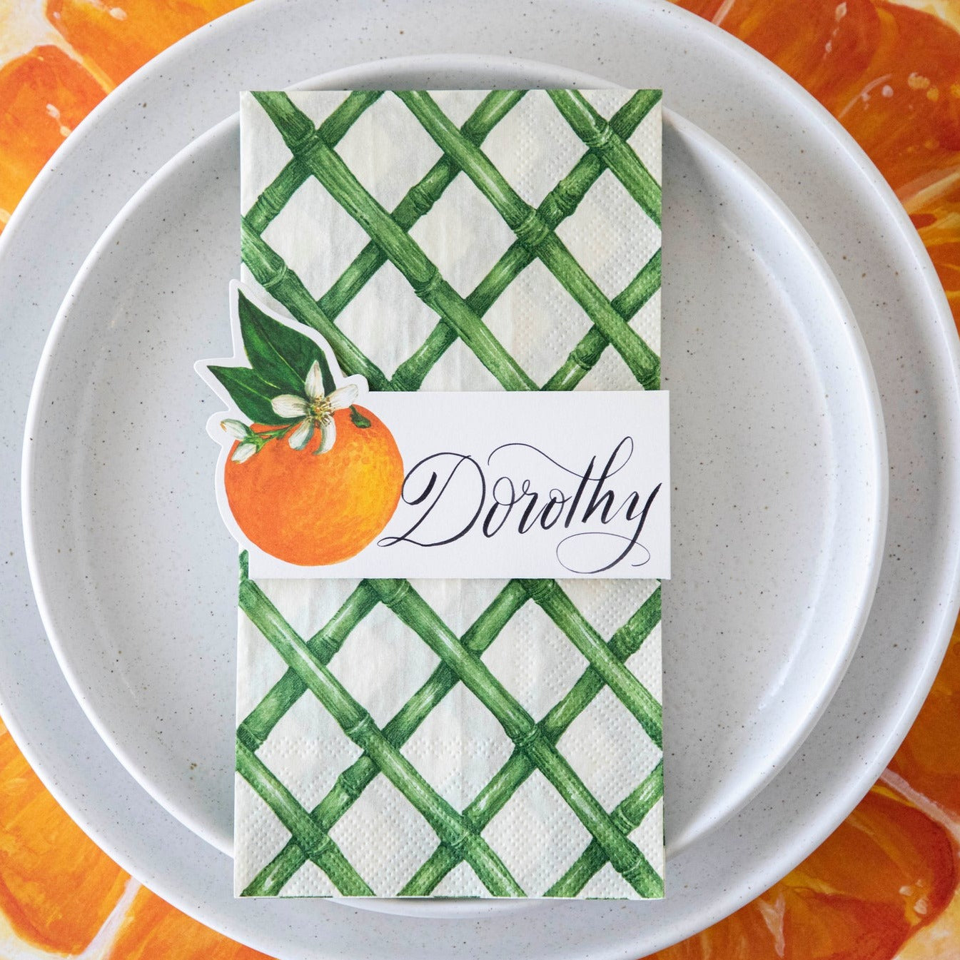 Top-down view of an citrus-themed place setting featuring an Orange Orchard Place Card labeled "Dorothy" laying flat on the Green Lattice Guest Napkin.