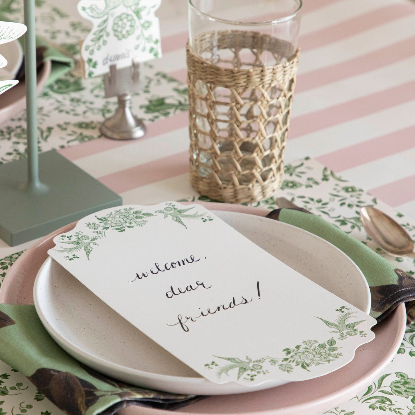 A Green Asiatic Pheasants Table Card with "Welcome, dear friends!" written on it in cursive resting on the plate of an elegant place setting.