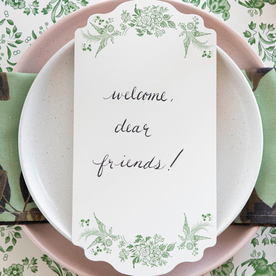 Close-up of a Green Asiatic Pheasants Table Card with &quot;Welcome, dear friends!&quot; written on it in cursive resting on the plate of an elegant place setting.
