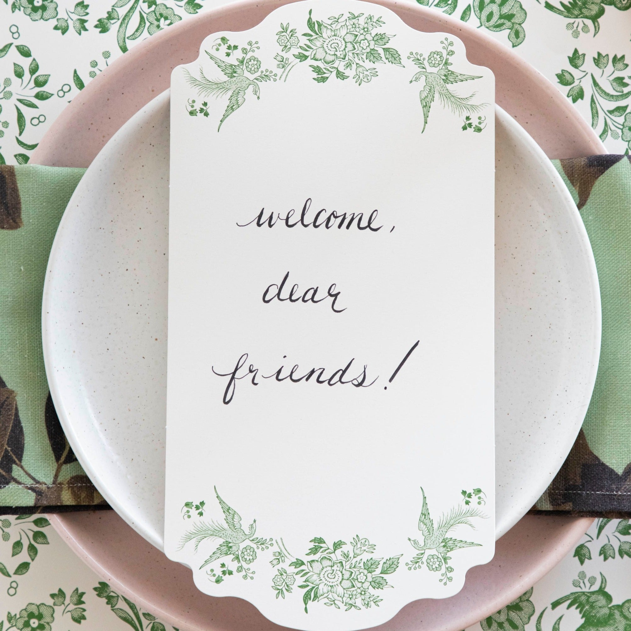 Close-up of a Green Asiatic Pheasants Table Card with "Welcome, dear friends!" written on it in cursive resting on the plate of an elegant place setting.