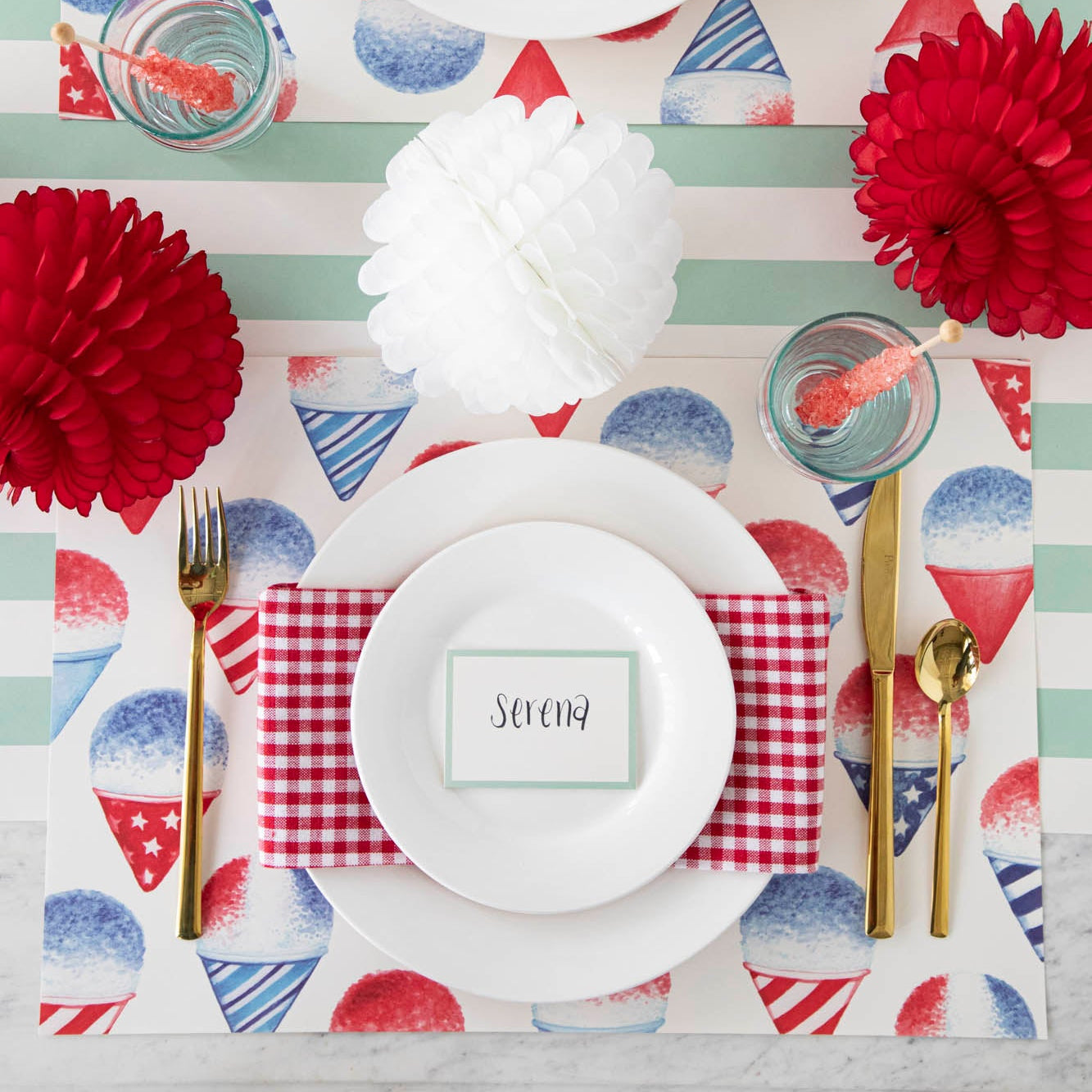 A patriotic place setting featuring the Snow Cone Placemat with white plates, a red-and-white checkered napkin, Seafoam Place Card reading "Serena" and gold flatware atop, on the Seafoam Classic Stripe Runner.