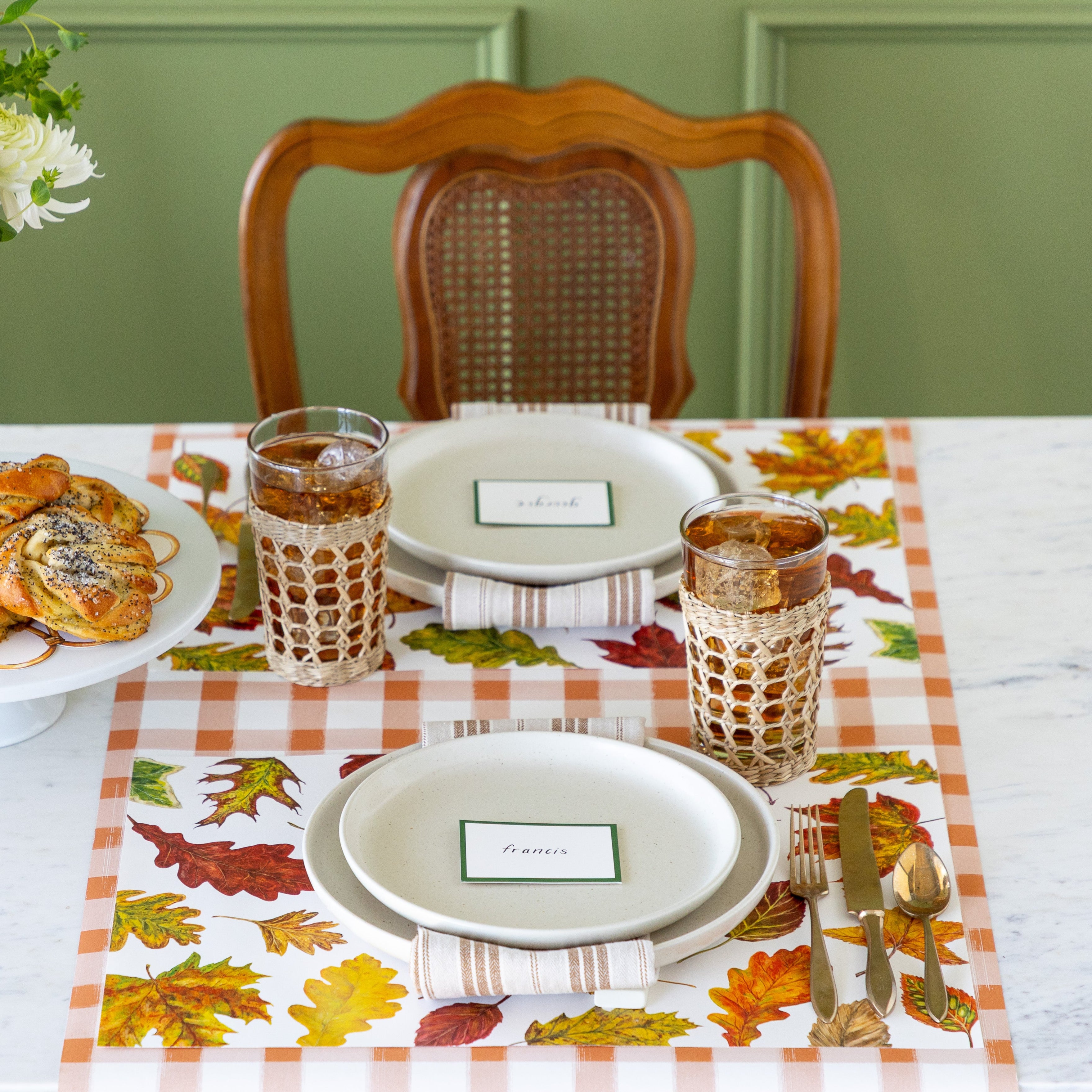An elegant table setting for two featuring the Fall Foliage Placemat under vintage flatware, dinner plates with the Dark Green Frame Place Card on top and a glass of iced tea, next to a cake stand with decadent pastries and flowers, and Orange Painted Check Runner running vertically down the table.