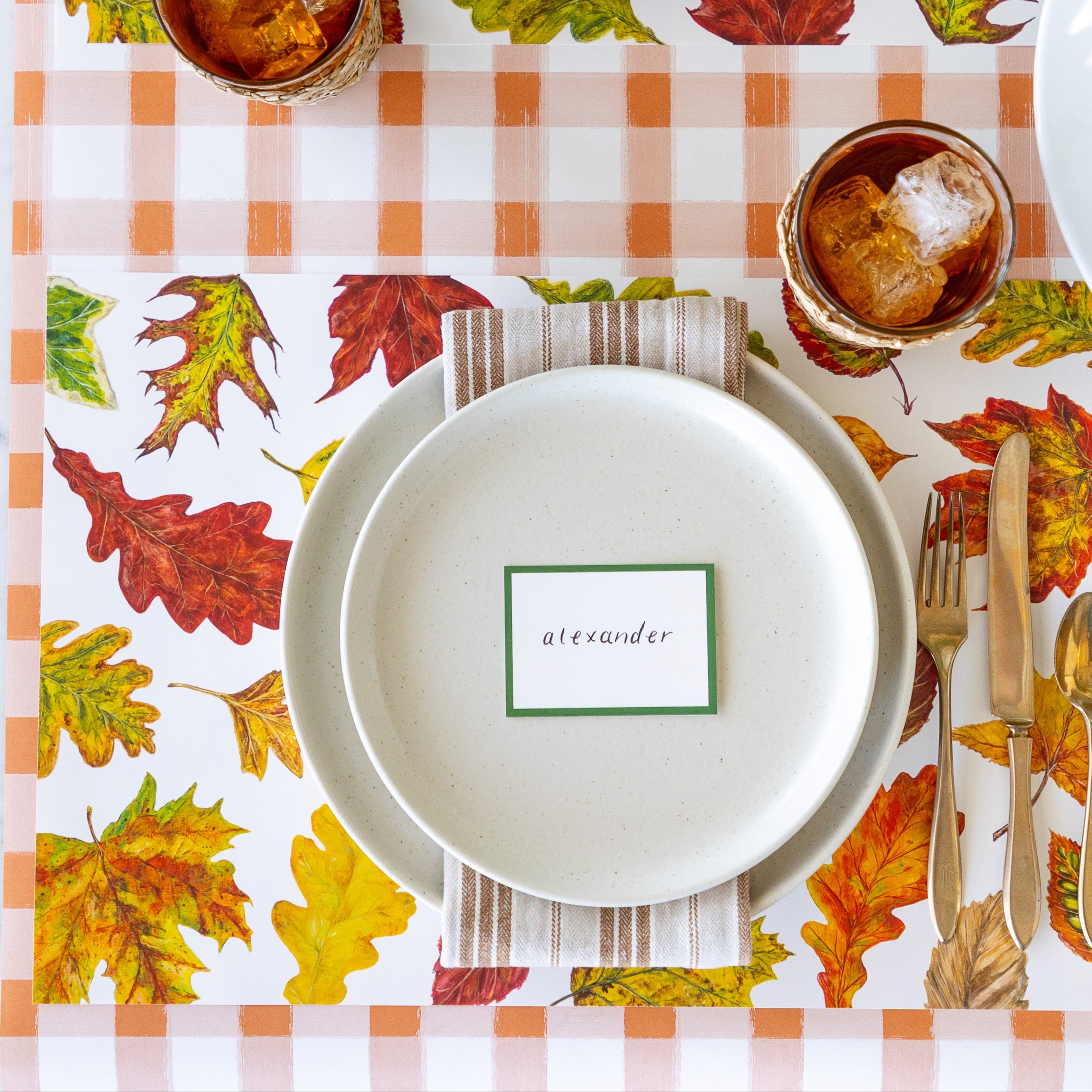 An elegant place setting featuring the Fall Foliage Placemat with a brown striped napkin in between two dinner plates with the Dark Green Frame Place Card reading "alexander", on top, a glass of iced tea and Orange Painted Check Runner underneath.