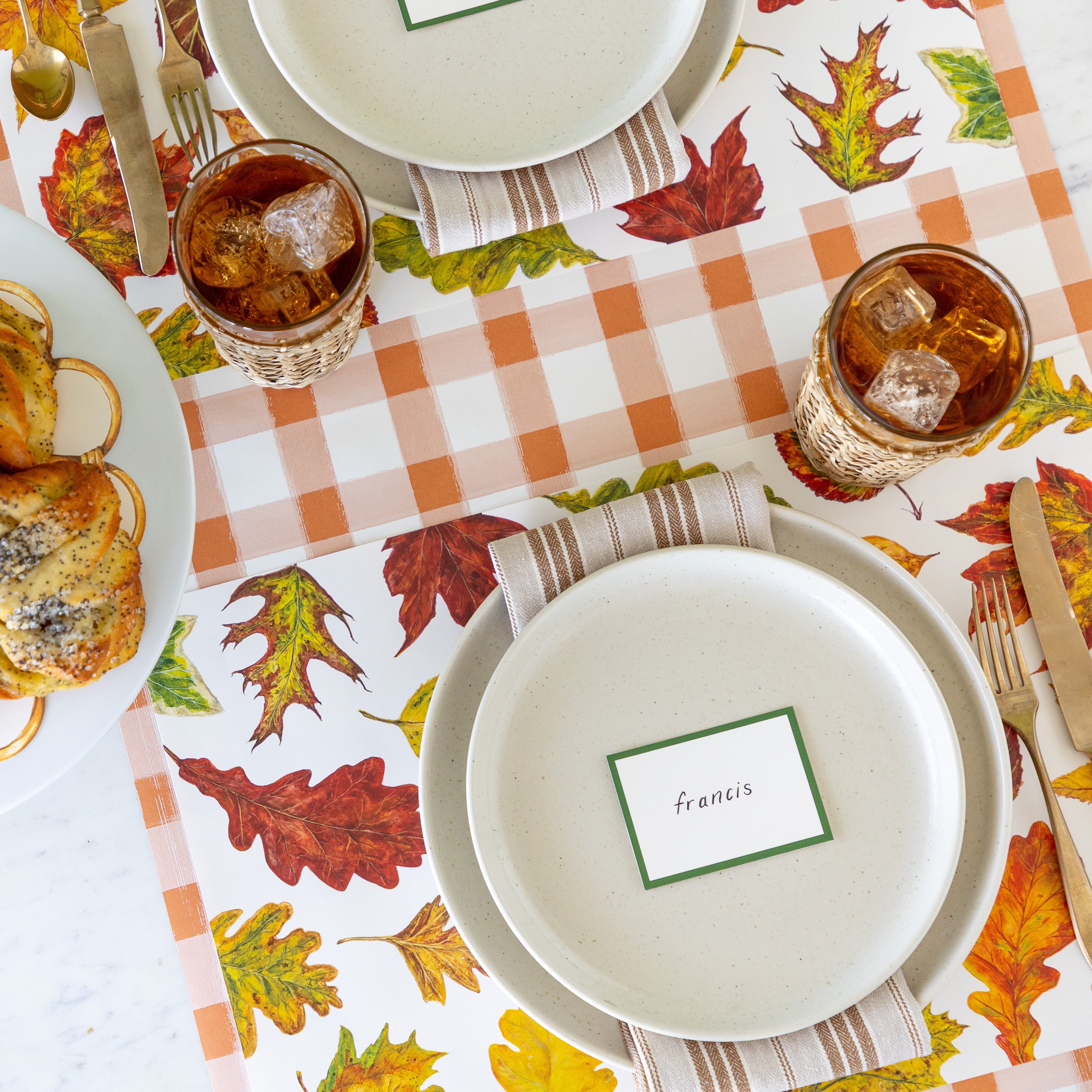 An elegant place setting featuring the Fall Foliage Placemat with a brown striped napkin in between two dinner plates with the Dark Green Frame Place Card reading "francis", on top, a glass of iced tea and Orange Painted Check Runner underneath.