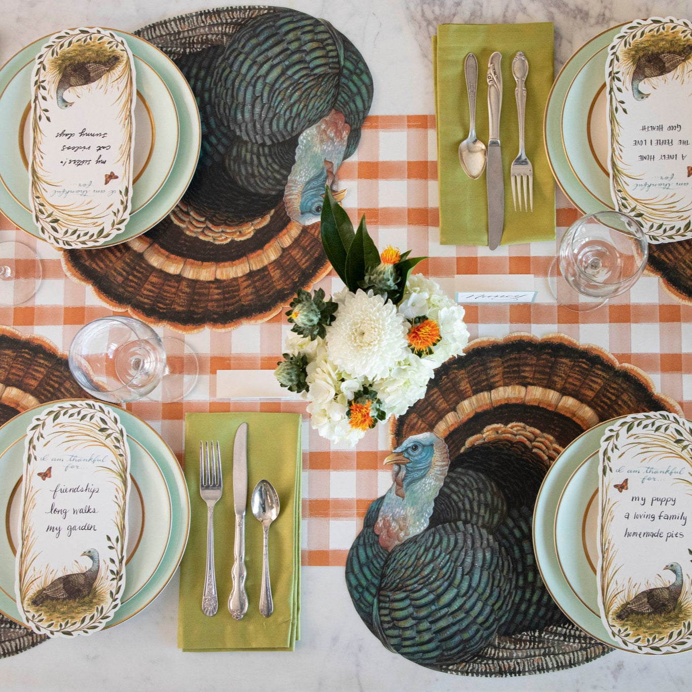 A turkey themed table setting featuring the Orange Painted Check Runner under Die-cut Heritage Turkey Placemats, seafoam dinner plates, Thankful Turkey Table Accents, green napkins with silver flatware atop, and a floral centerpiece.