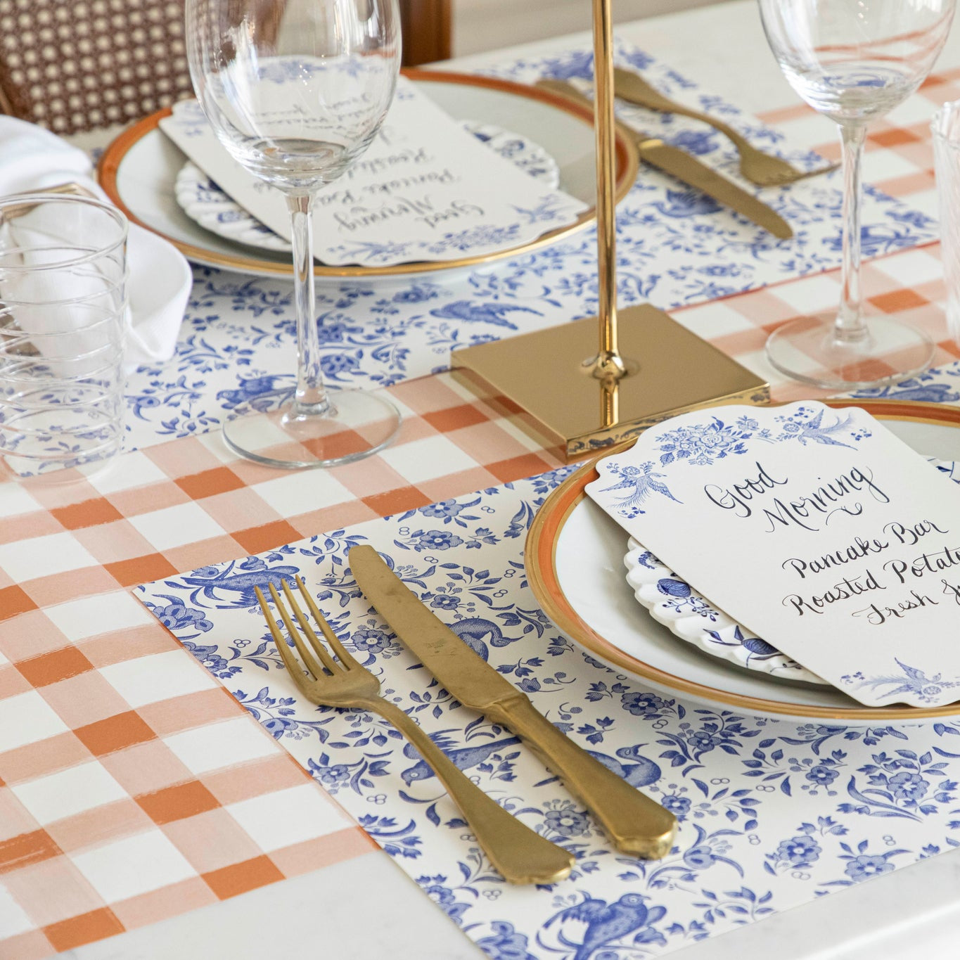 An elegant table setting featuring the Orange Painted Check Runner under the Burleigh Blue Regal Peacock Placemats, vintage dinnerware with Blue Regal Peacock Table Cards atop, gold flatware and wine glasses.
