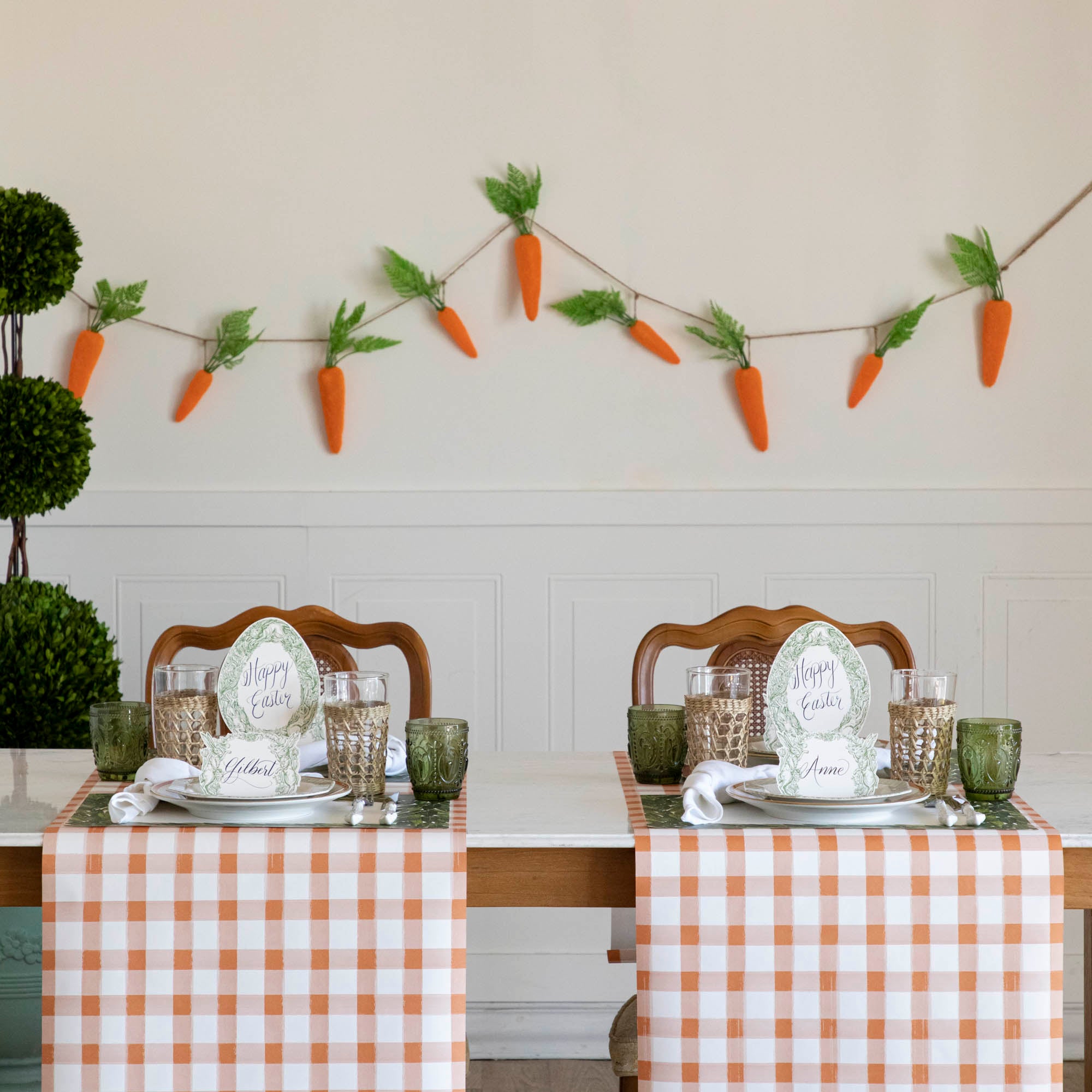 An Easter themed dining room featuring the Orange Painted Check Runners, green and seagrass cage glasses, a carrot garland hanging on the wall and a boxwood topiary.
