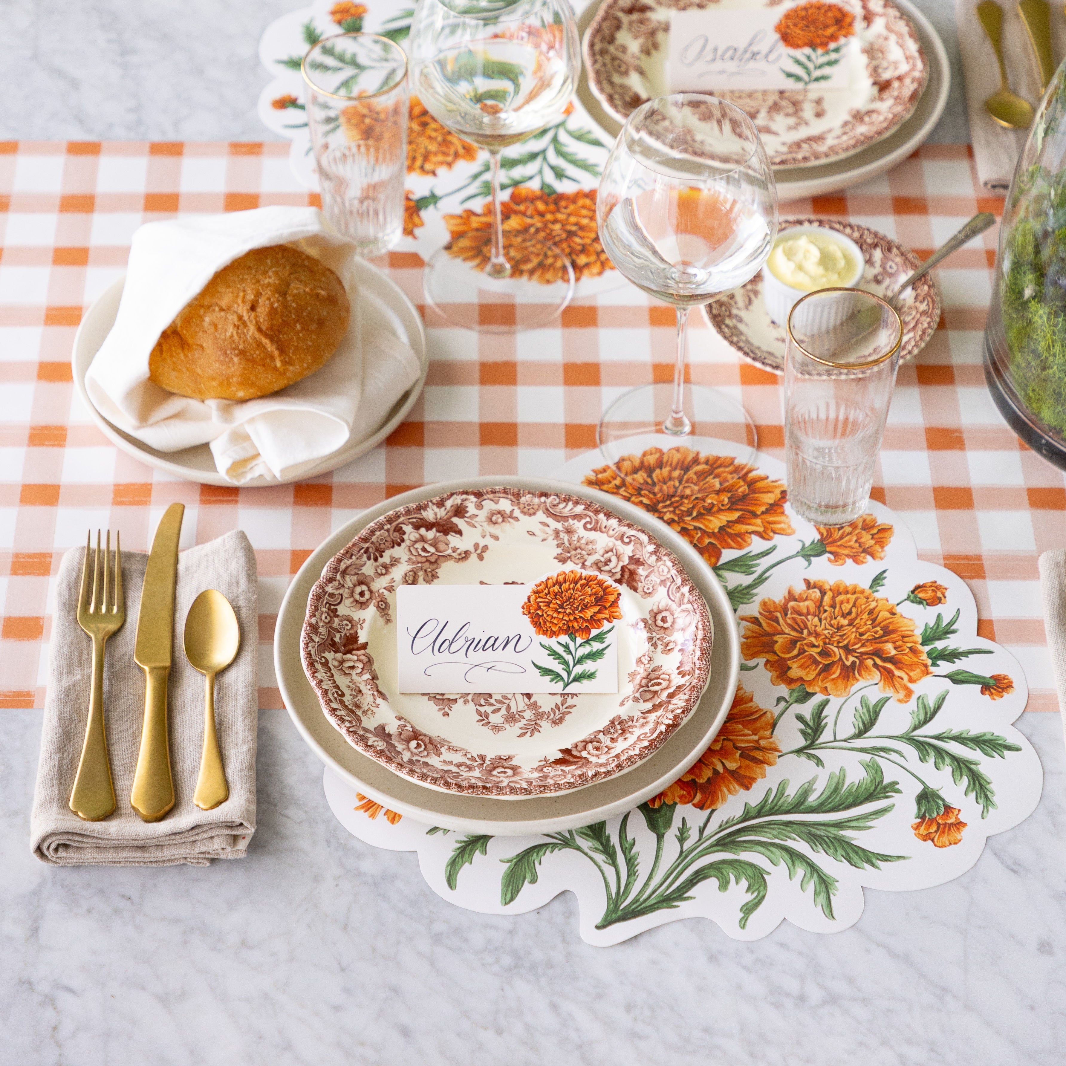 An elegant table setting featuring the Die-cut Marigold Bouquet Placemat offset under vintage floral dinnerware with the Marigold Bloom Place Card on top, a wine and water glass, Mepra Gold Flatware on a napkin nearby, and the Orange Painted Check Runner underneath.