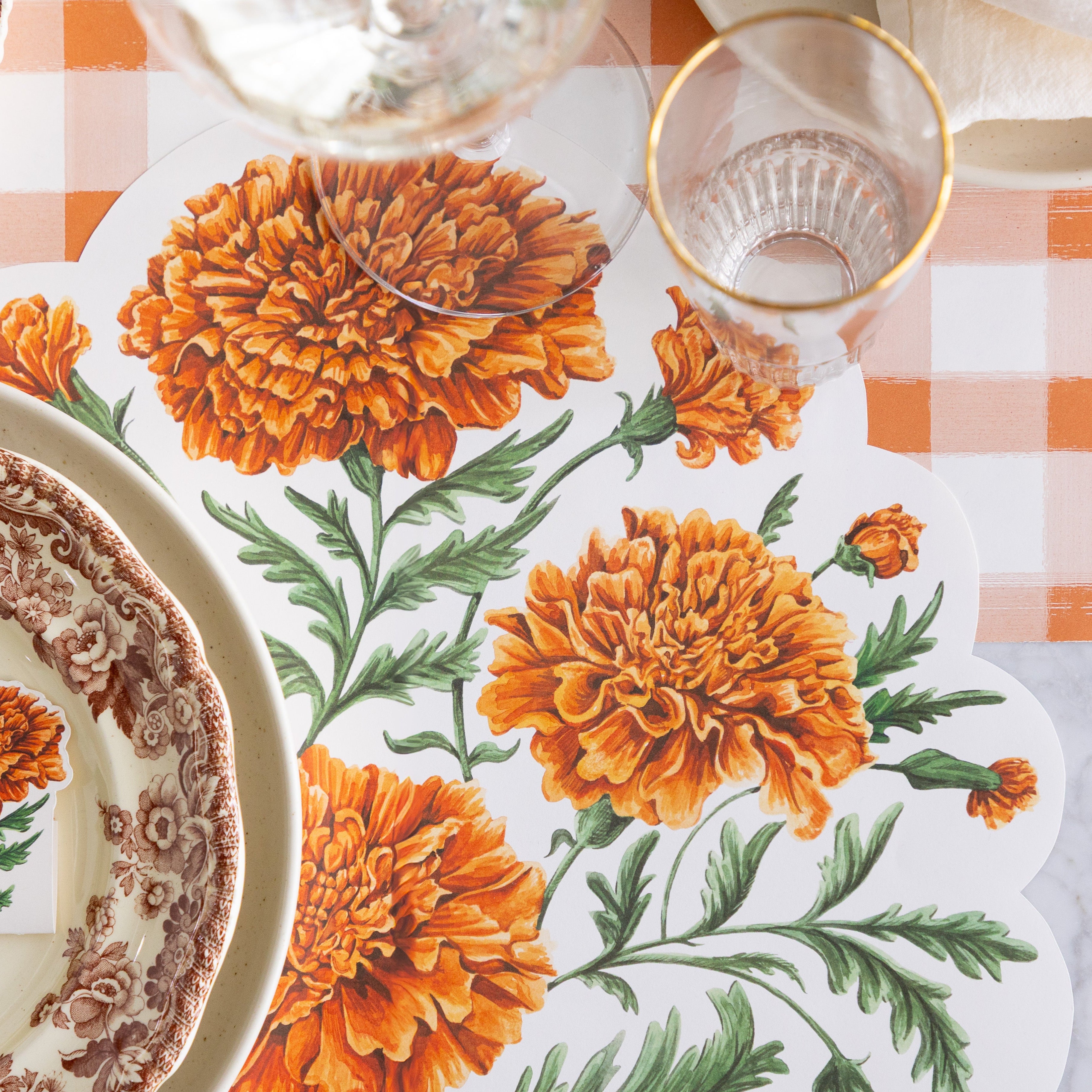 An elegant place setting featuring the Die-cut Marigold Bouquet Placemat offset to the right of the dinner plates, with a wine and water glass on top and Orange Painted Check Runner underneath.
