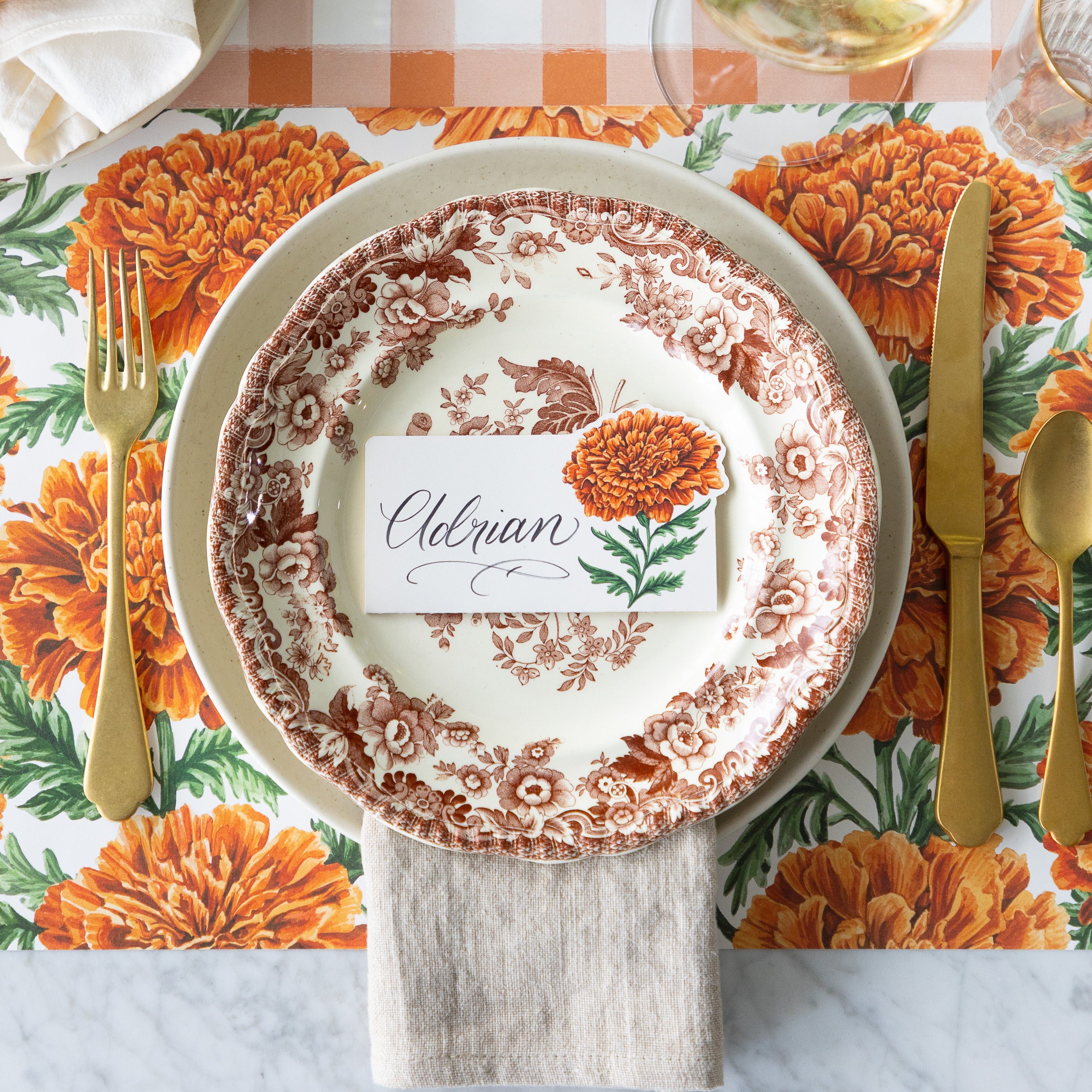 A place setting featuring the Marigold Bloom Place Card reading "Adrian" atop vintage dinner plates with the Marigold Harvest Placemat and Orange Painted check Runner underneath.