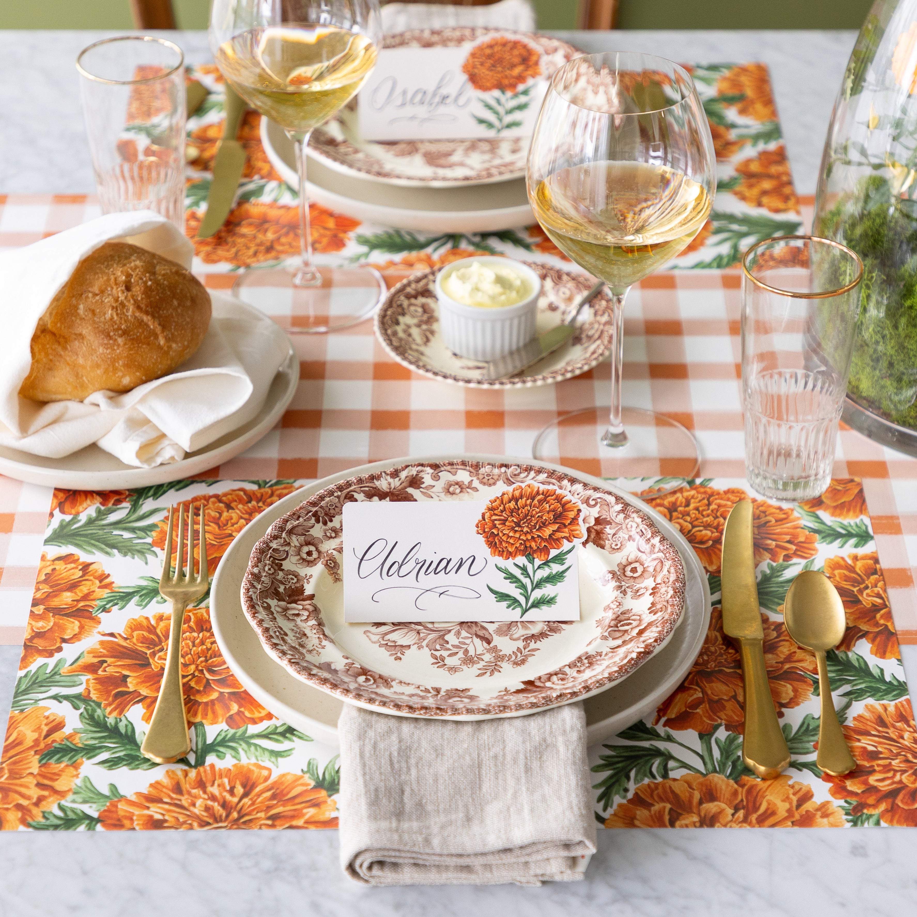 An elegant table setting with a vintage floral plate, gold utensils, bread roll, wine glass, and a Marigold Bloom Place Card reading "Eleanor" atop a Marigold Harvest Placemat—on a Orange Painted Check Runner.