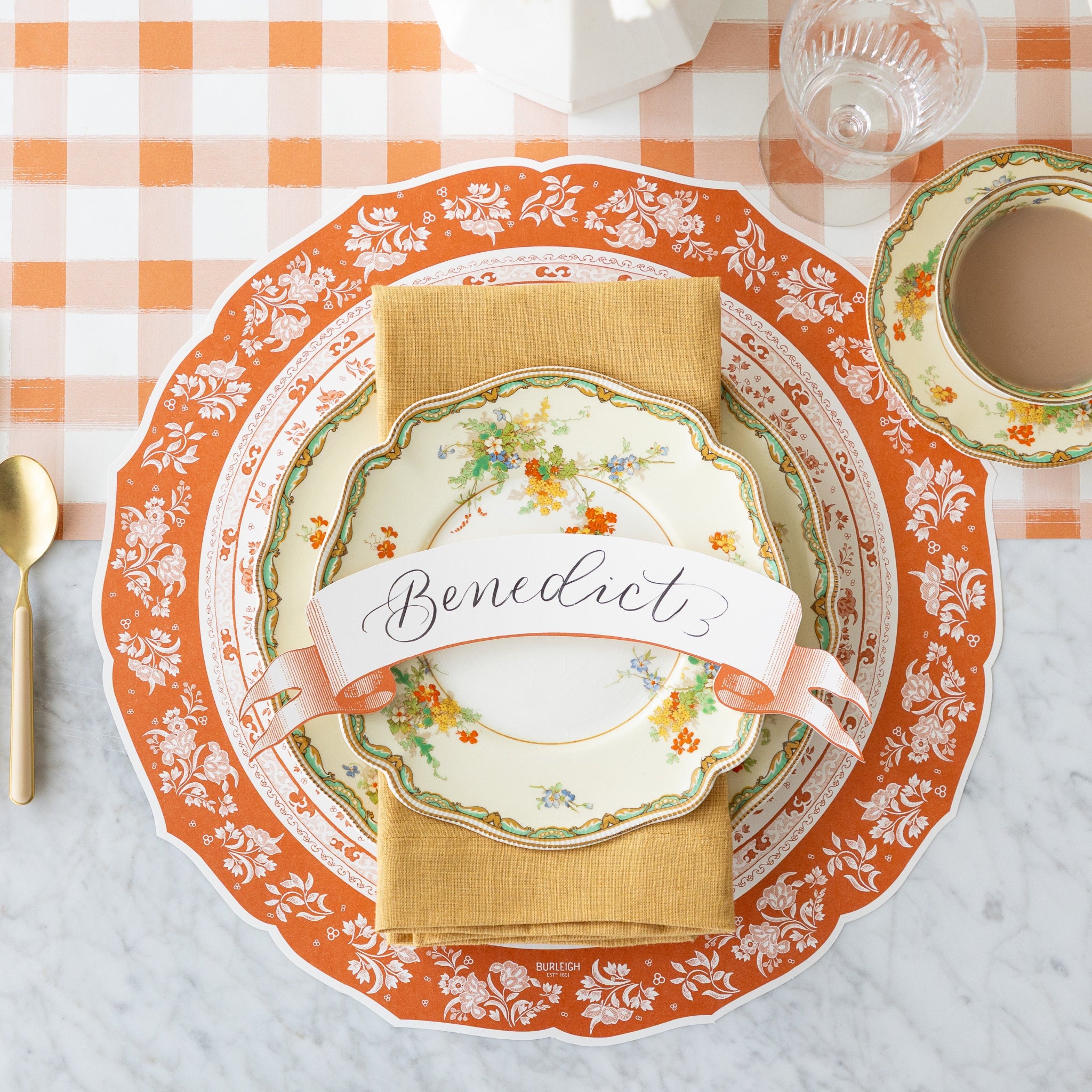 A fall place setting featuring the Die-cut Pumpkin Regal Placemat, vintage floral dinnerware with a tan napkin and Orange Banner Table Accent reading "Benedict", atop, and a cup of coffee on the Orange Painted Check Runner.