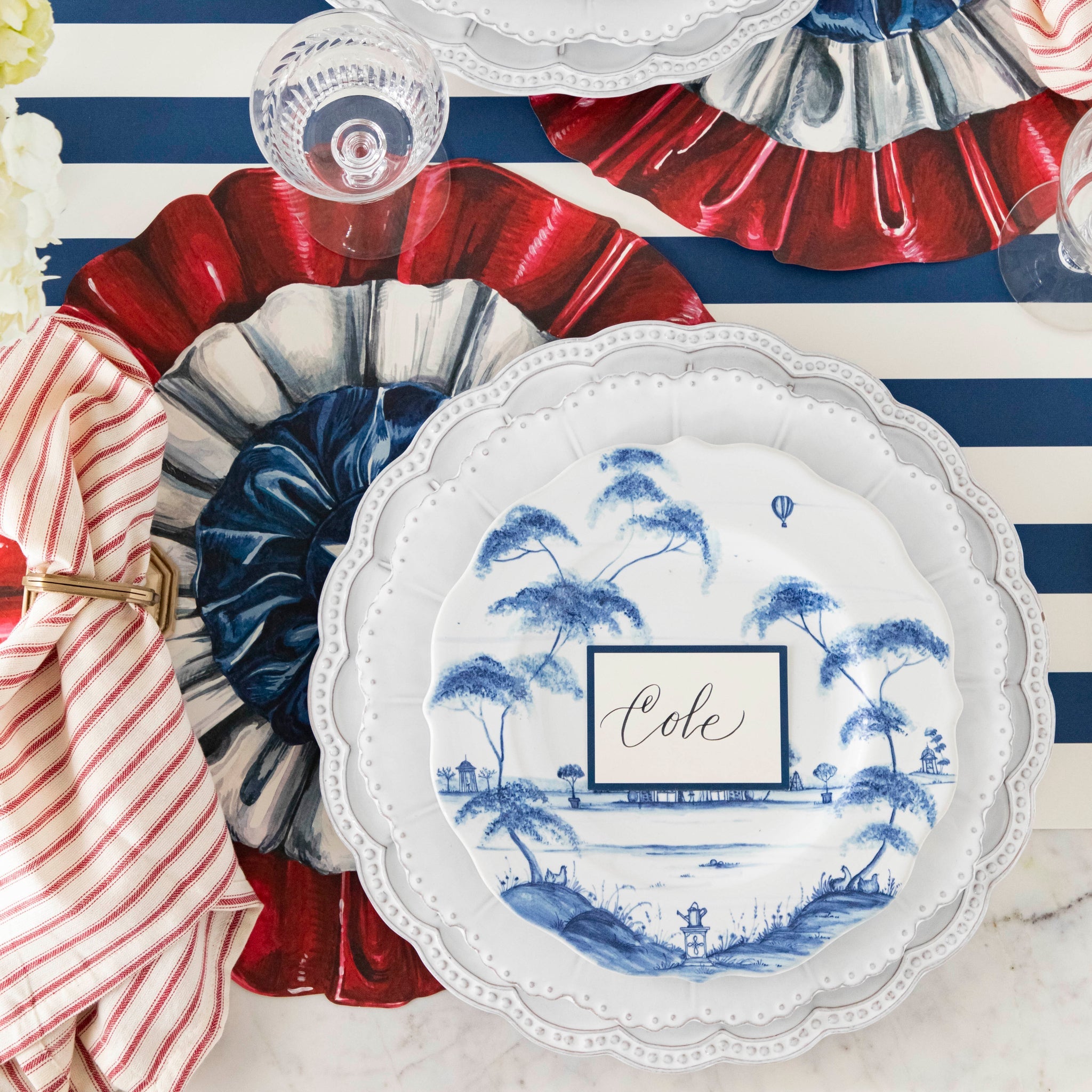 Patriotic table setting featuring the Die-cut Star Spangled Placemat, a red-and-white striped napkin, beaded and blue floral plates, and Blue Frame Place Card on the Navy Classic Stripe Runner.