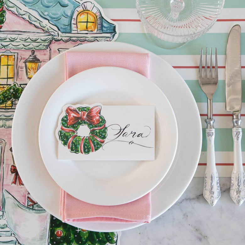 A festive holiday place setting with a Holiday Wreath Place Card labeled "Sara" laying flat on the plate, from above.