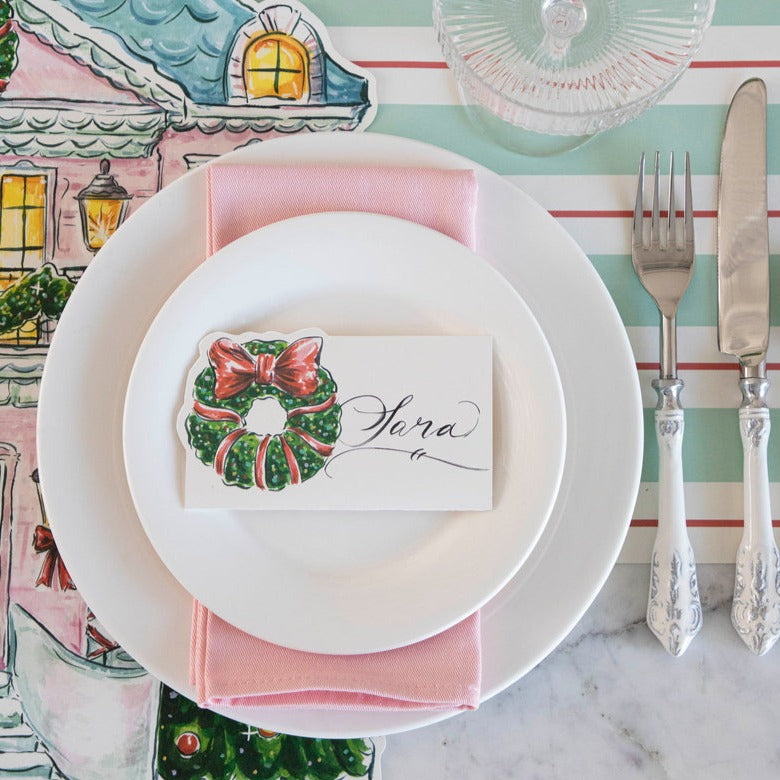 A festive holiday place setting with a Holiday Wreath Place Card labeled &quot;Sara&quot; laying flat on the plate, from above.