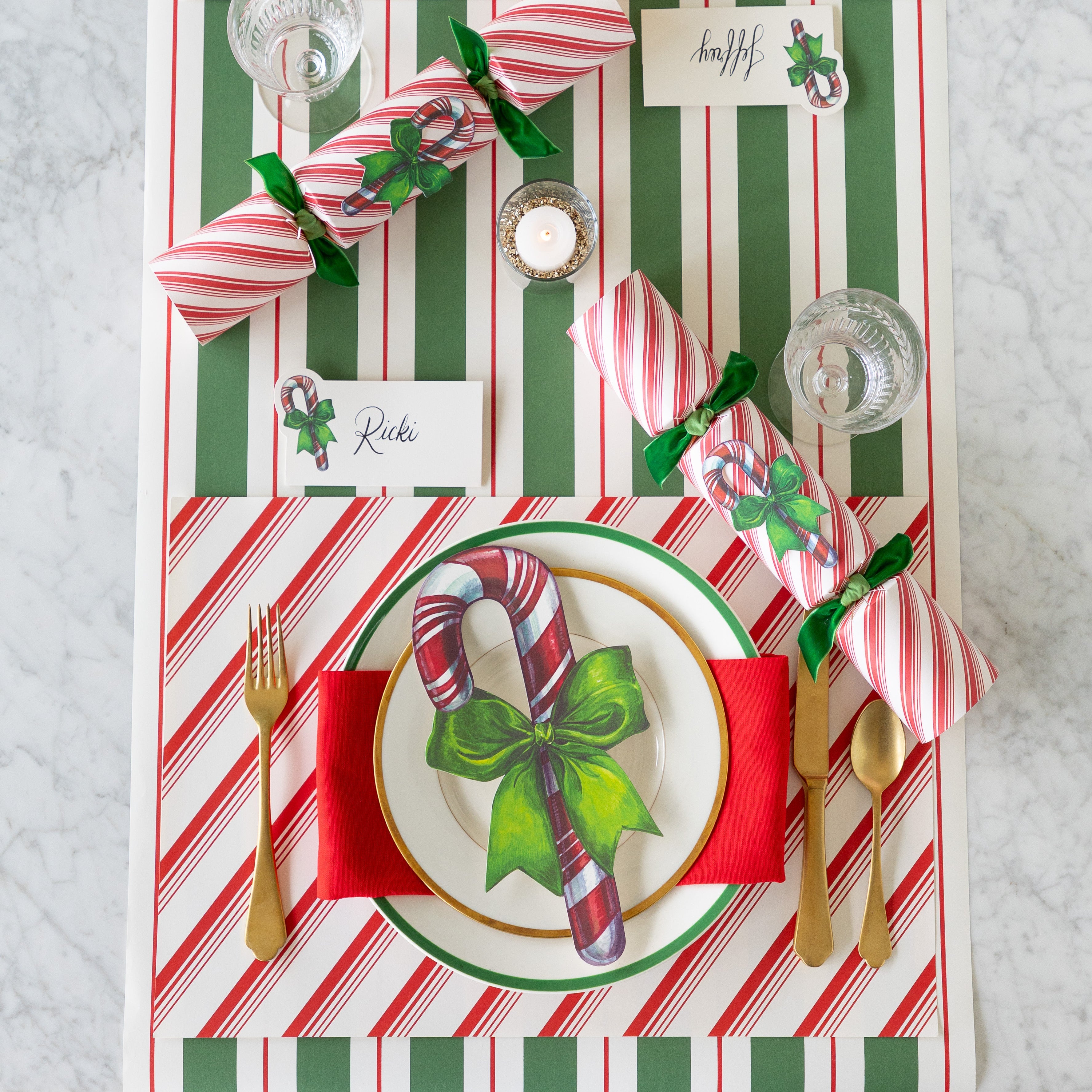 A place setting featuring the Peppermint Stripe Placemat, gold flatware, gold and green trimmed plates with the Candy Cane Table Accent atop, Candy Candy Place Card, Candy Cane Crackers and Green & Red Awning Stripe Runner underneath.