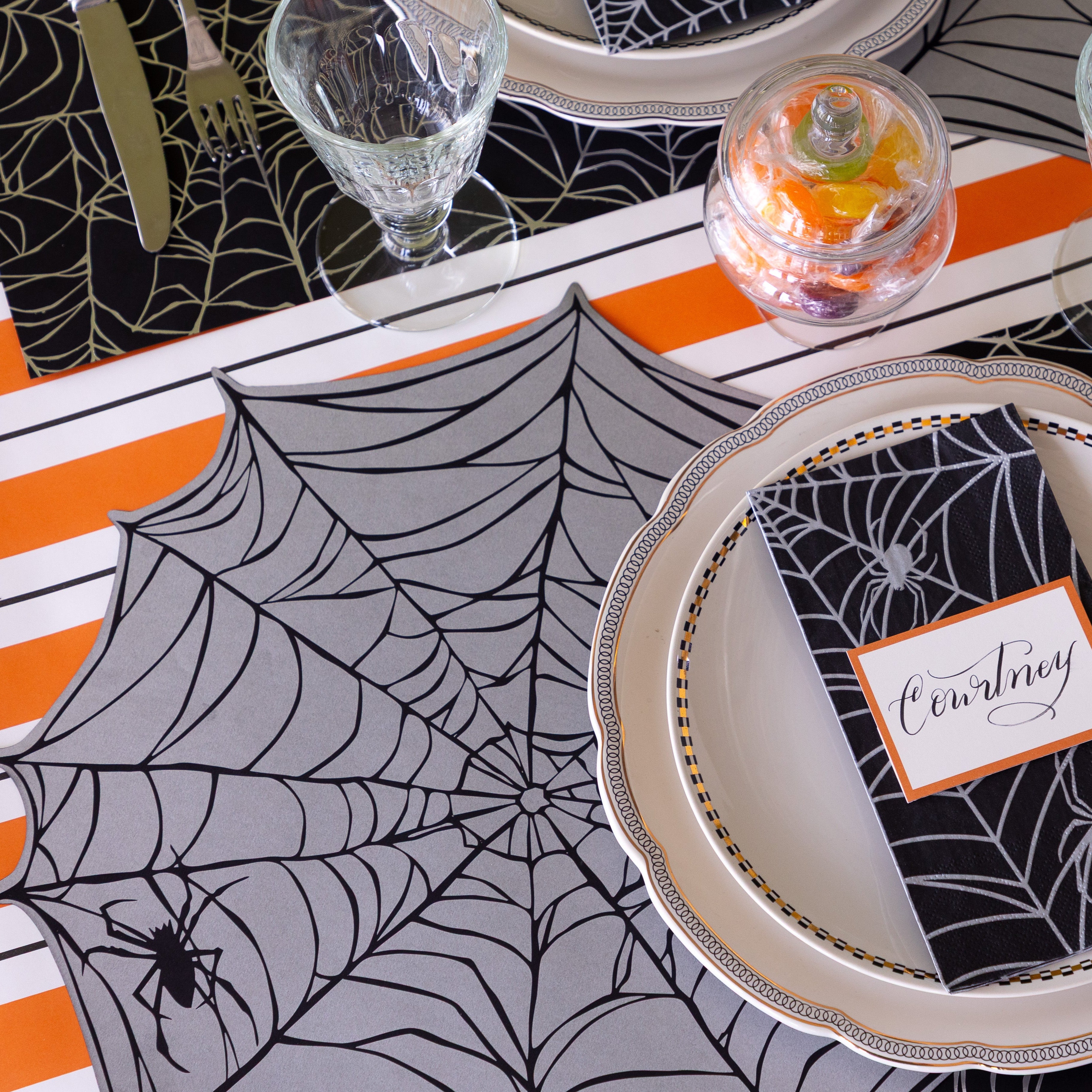 A Halloween themed table setting featuring the Die-cut Spiderweb Placemat, Spiderweb Guest Napkin on the plates with the Orange Frame Place Card on top, and Orange & Black Awning Stripe Runner underneath.