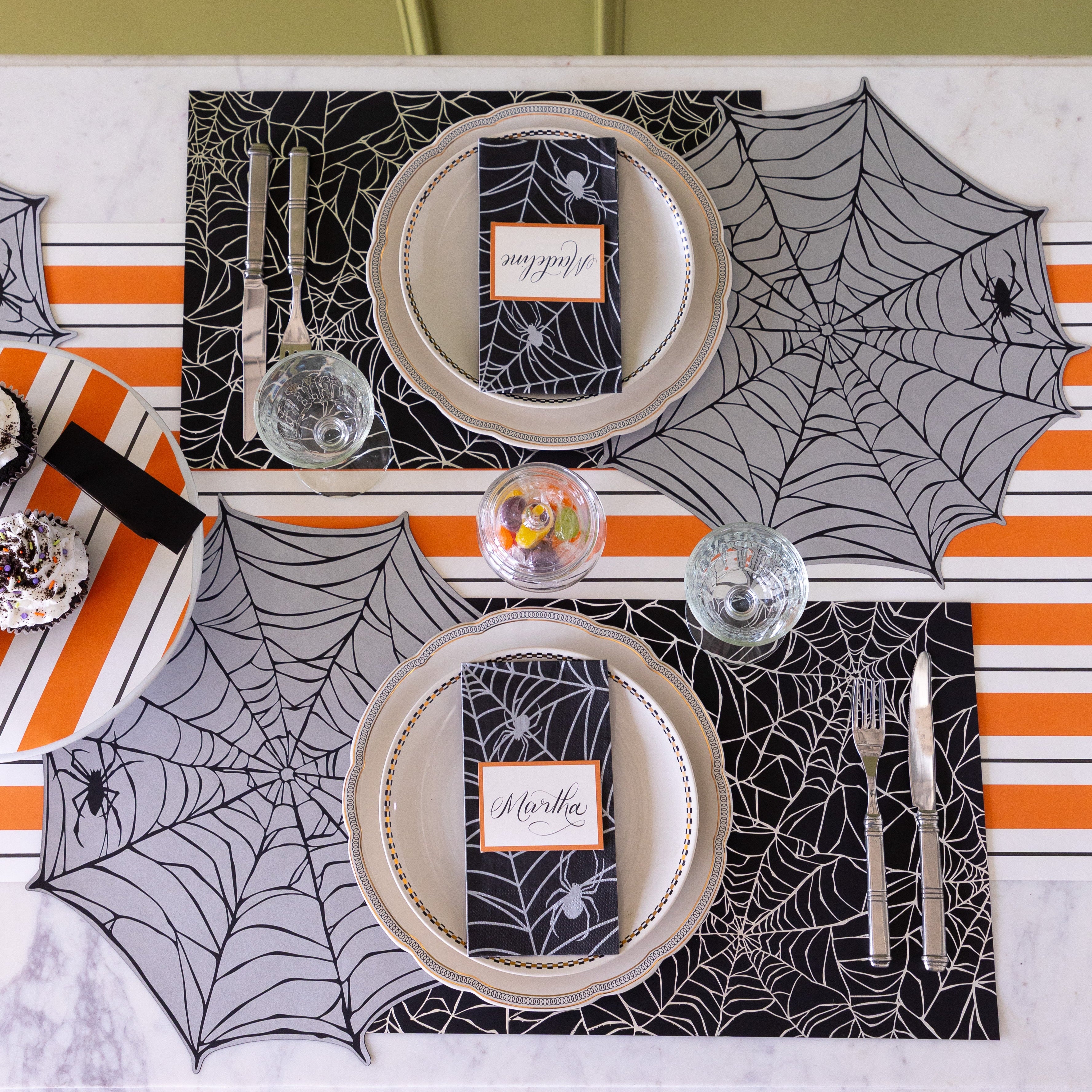 A Halloween themed table setting with candy and cupcakes, featuring the Die-cut Spiderweb Placemat slightly overlapping the Spiderweb Placemat, the Spiderweb Guest Napkin with Orange Frame Place Card on top and Orange & Black Awning Stripe Runner underneath.