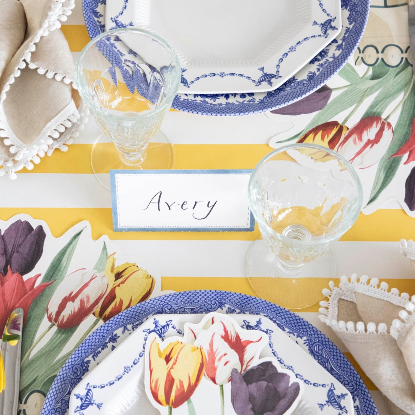 A table setting with tulips on a yellow and white striped tablecloth, framed by a Cornflower Blue Place Card from Hester &amp; Cook.