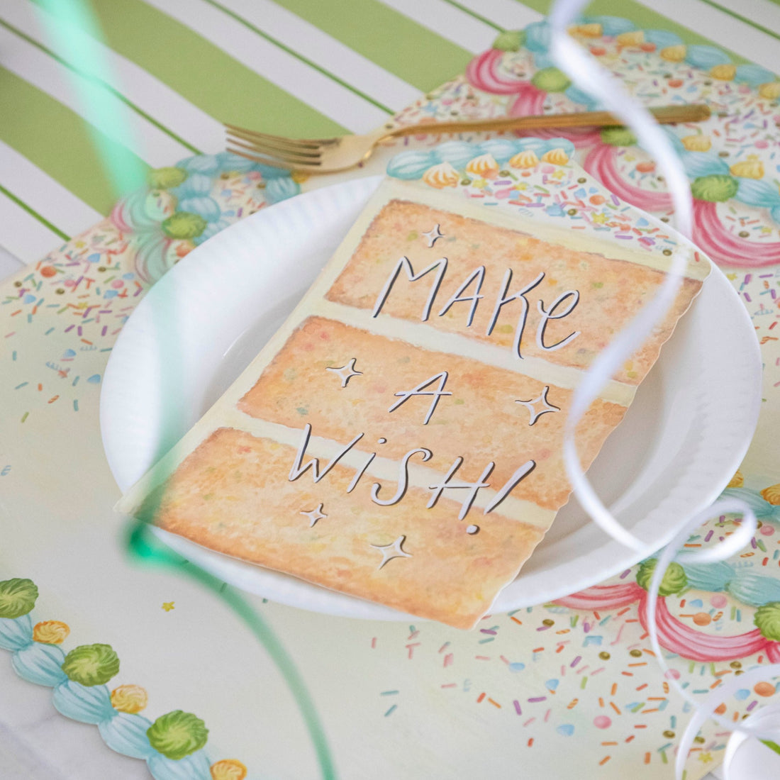 A vibrant birthday party place setting featuring a Cake Slice Table Accent resting on the plate with &quot;Make A Wish!&quot; written in white ink with a black outline.