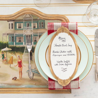 An Antique Gold Frame Table Card with a menu written on it in lovely cursive resting on the pate of an elegant place setting, from above.