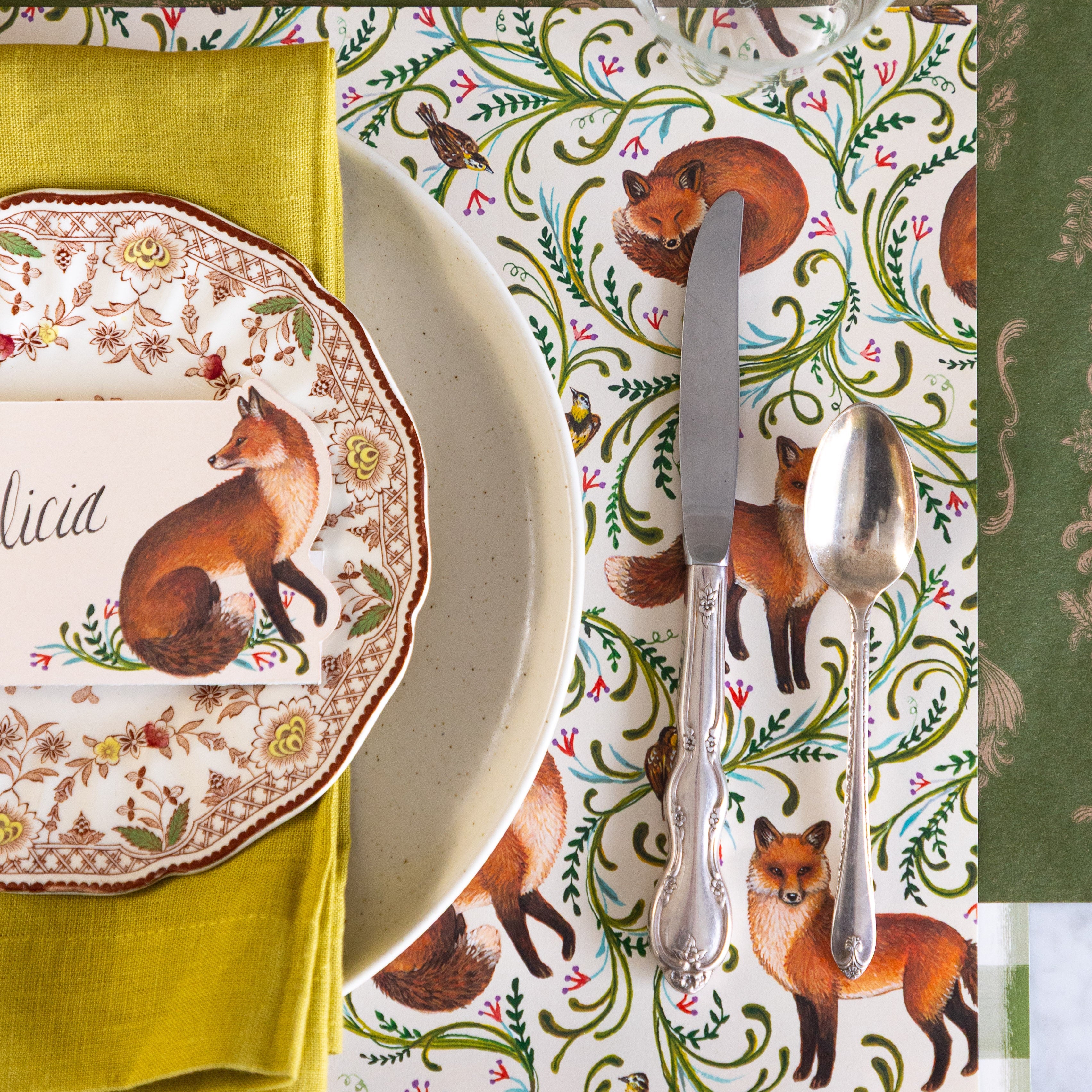 An elegant place setting featuring the Fanciful Foxes Placemat, vintage silverware, Pacifica Salt Dinner Plate with a green napkin, vintage floral plate and Fanciful Fox Place card on top, and Moss Fable Toile Runner under the place setting.