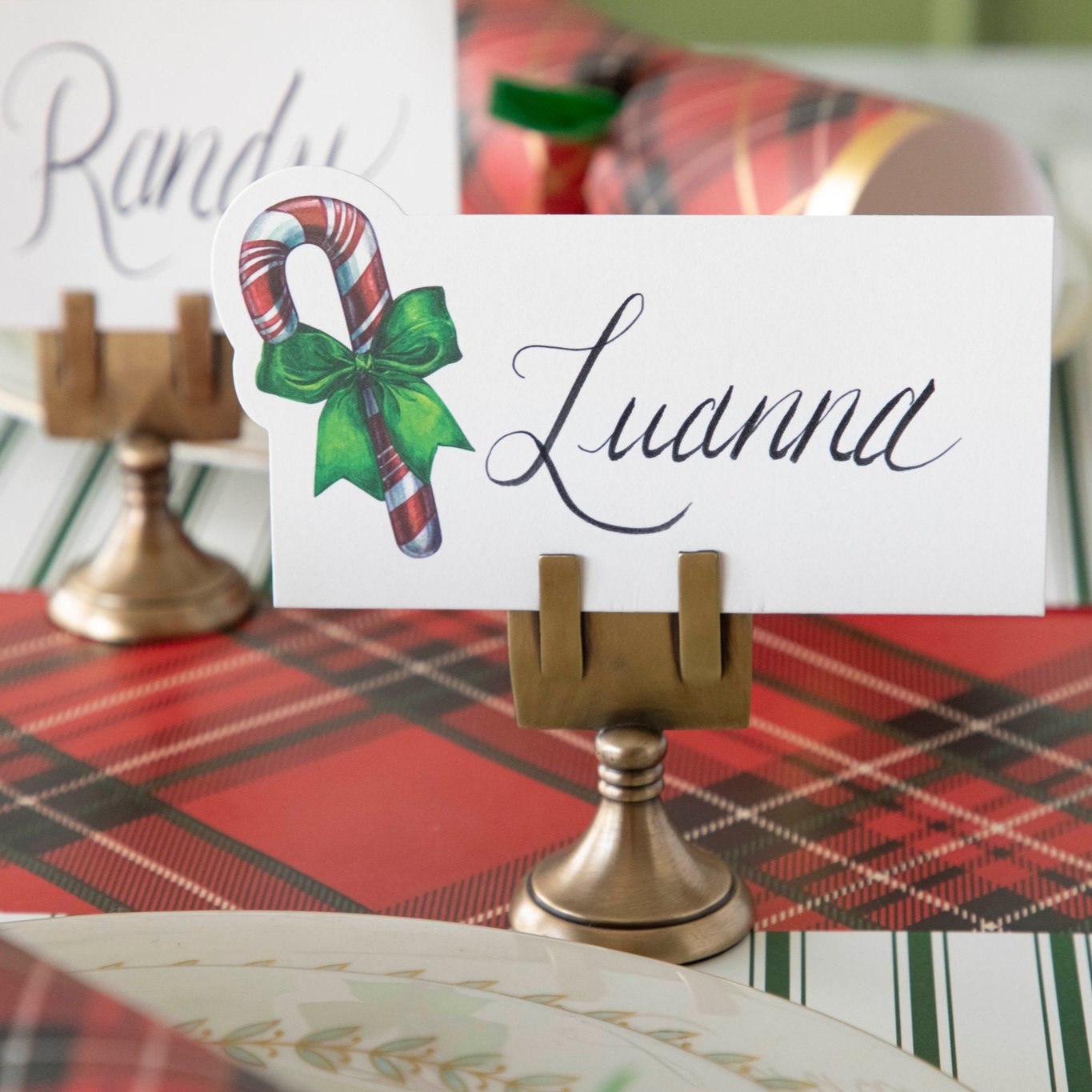 The Candy Cane Place Card with "Luanna" written on it, held up by the Brass Place Card Holder, on a festive table setting.