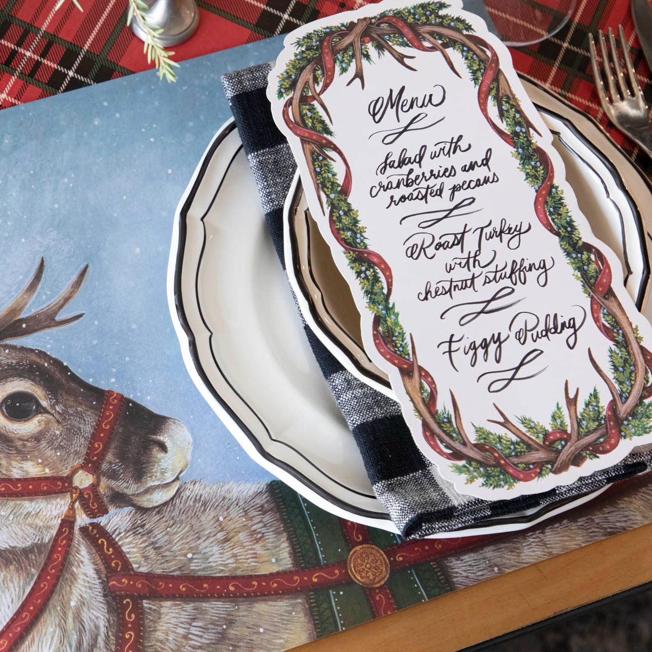 An Antler Garland Table Card with a menu written on it in lovely cursive, resting on the plate of a reindeer-themed Christmas place setting.