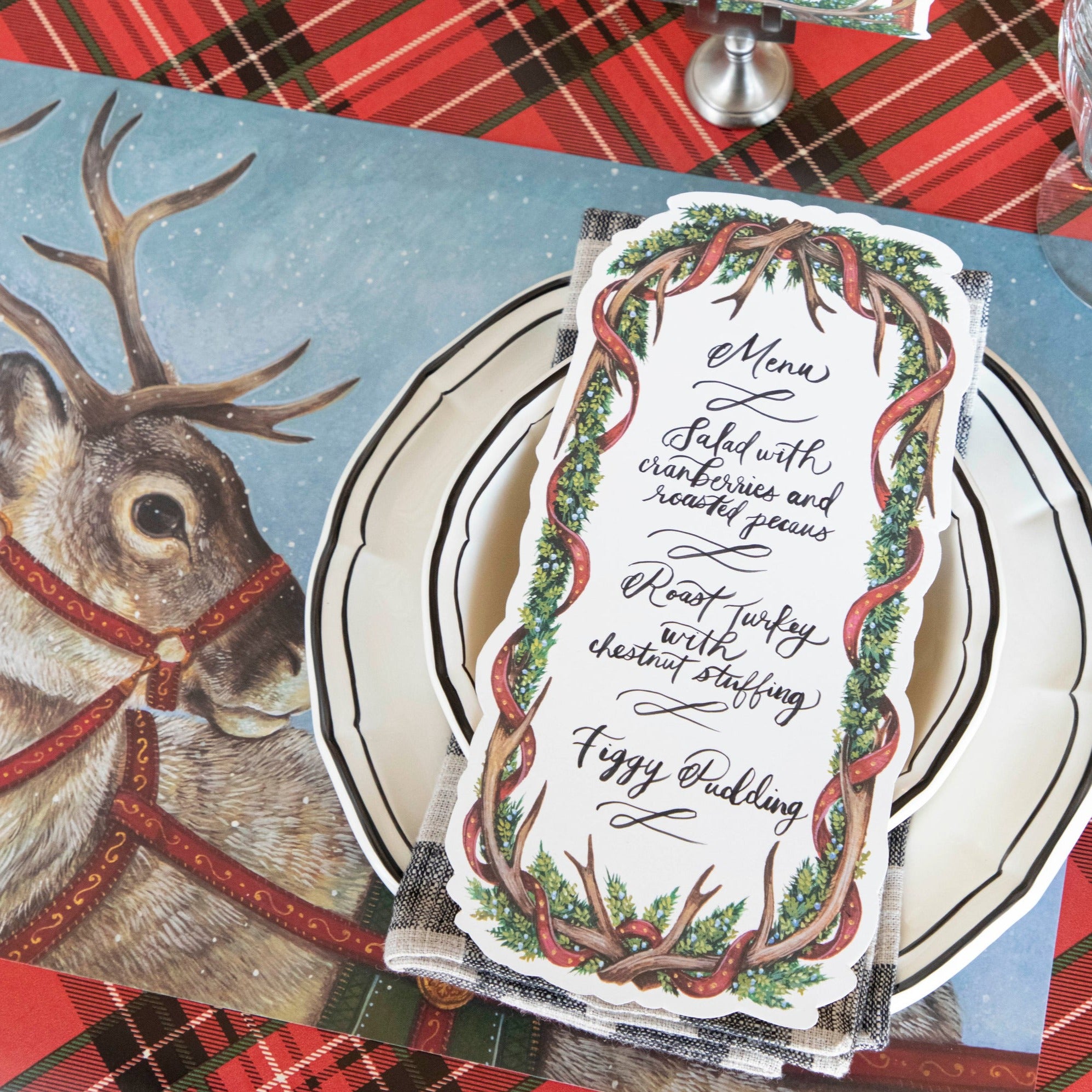 An Antler Garland Table Card with a menu written on it in lovely cursive, resting on the plate of a reindeer-themed Christmas place setting.