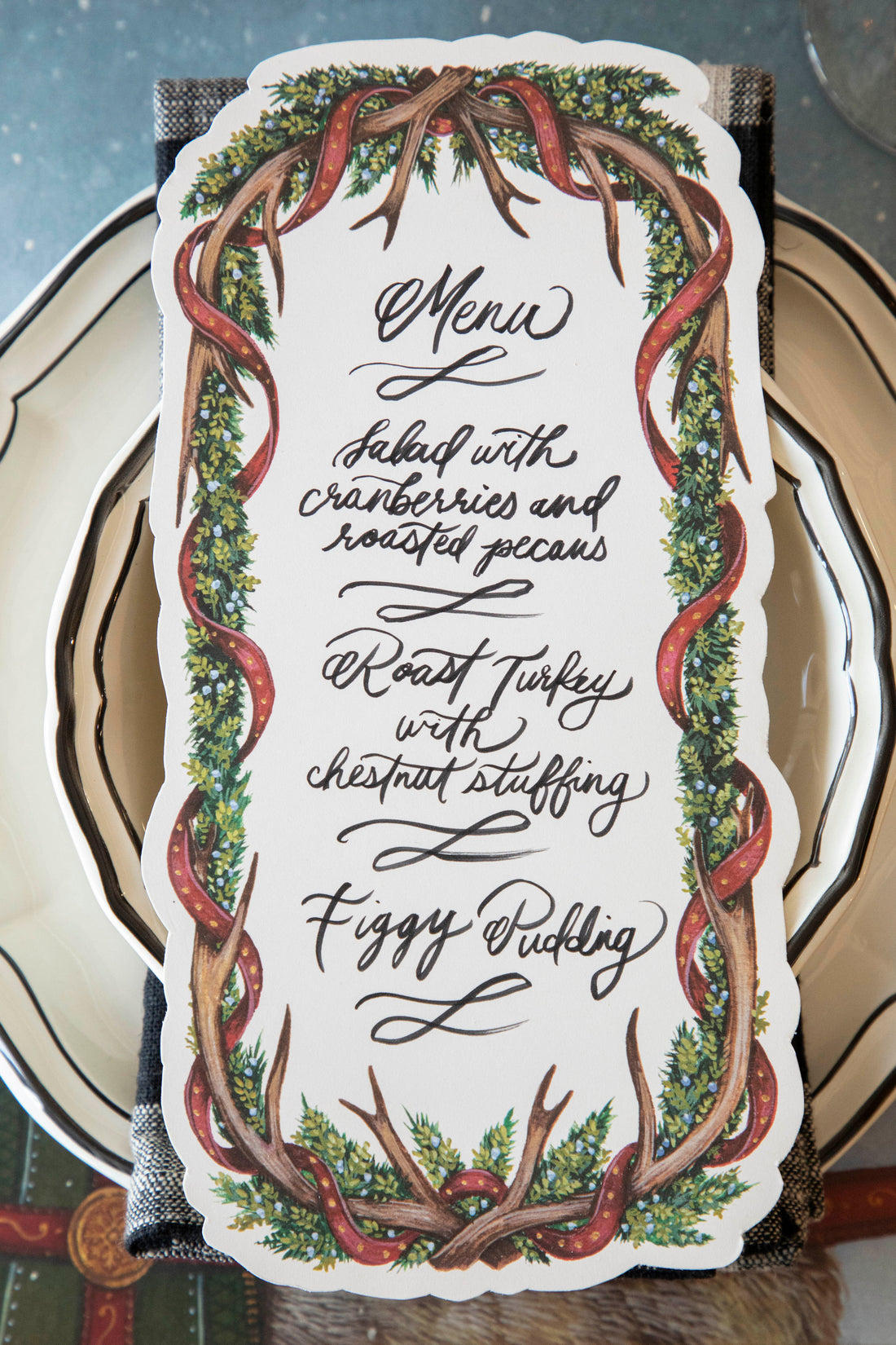 An Antler Garland Table Card with a menu written on it in lovely cursive, resting on the plate of a Christmas place setting.