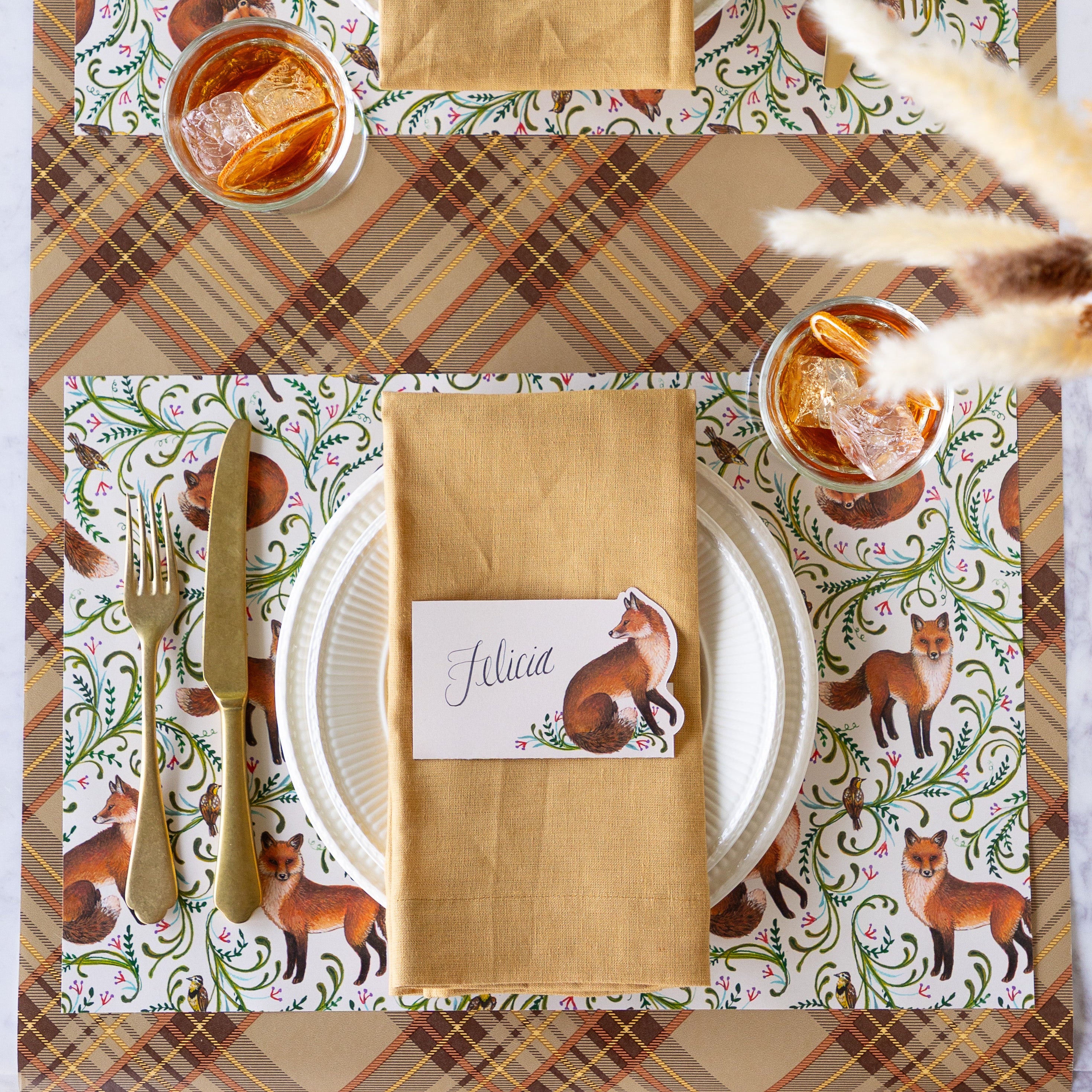An elegant place setting featuring the Fanciful Foxes Placemat, Mepra Gold fork and knife, white dinner plates with a tan napkin and Fanciful Fox Place card on top, and Autumn Plaid Runner underneath.