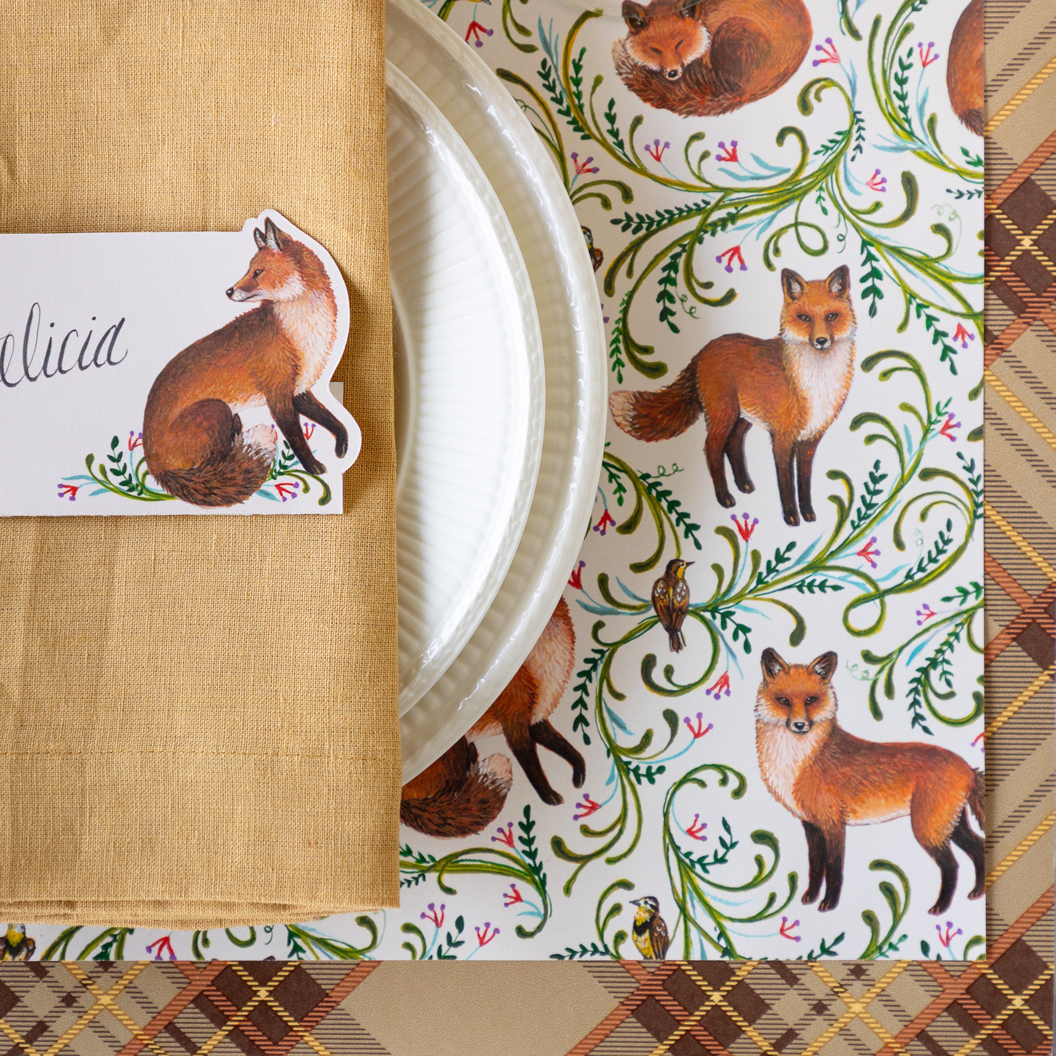 A close up of the Fanciful Fox Placemat on the Autumn Plaid Runner with white dinnerware, a tan napkin and Fanciful Fox Place Card on top.
