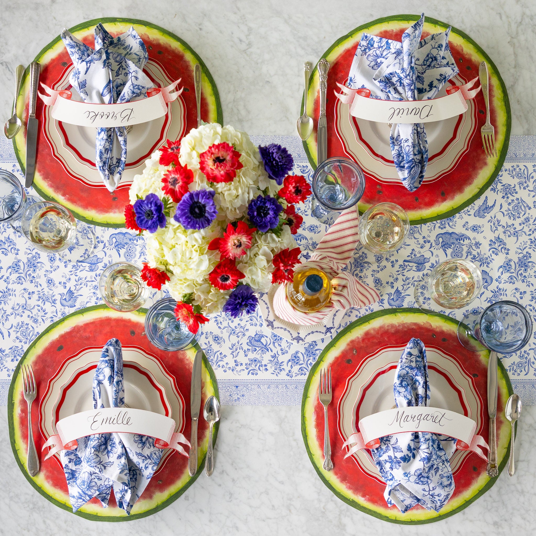 A summery table setting for four with a floral centerpiece, featuring the Die-cut Watermelon Placemats, red scalloped plates, blue floral napkins, Classic Red Banner Table Accents, and glasses of wine atop the Blue Regal Peacock Runner.