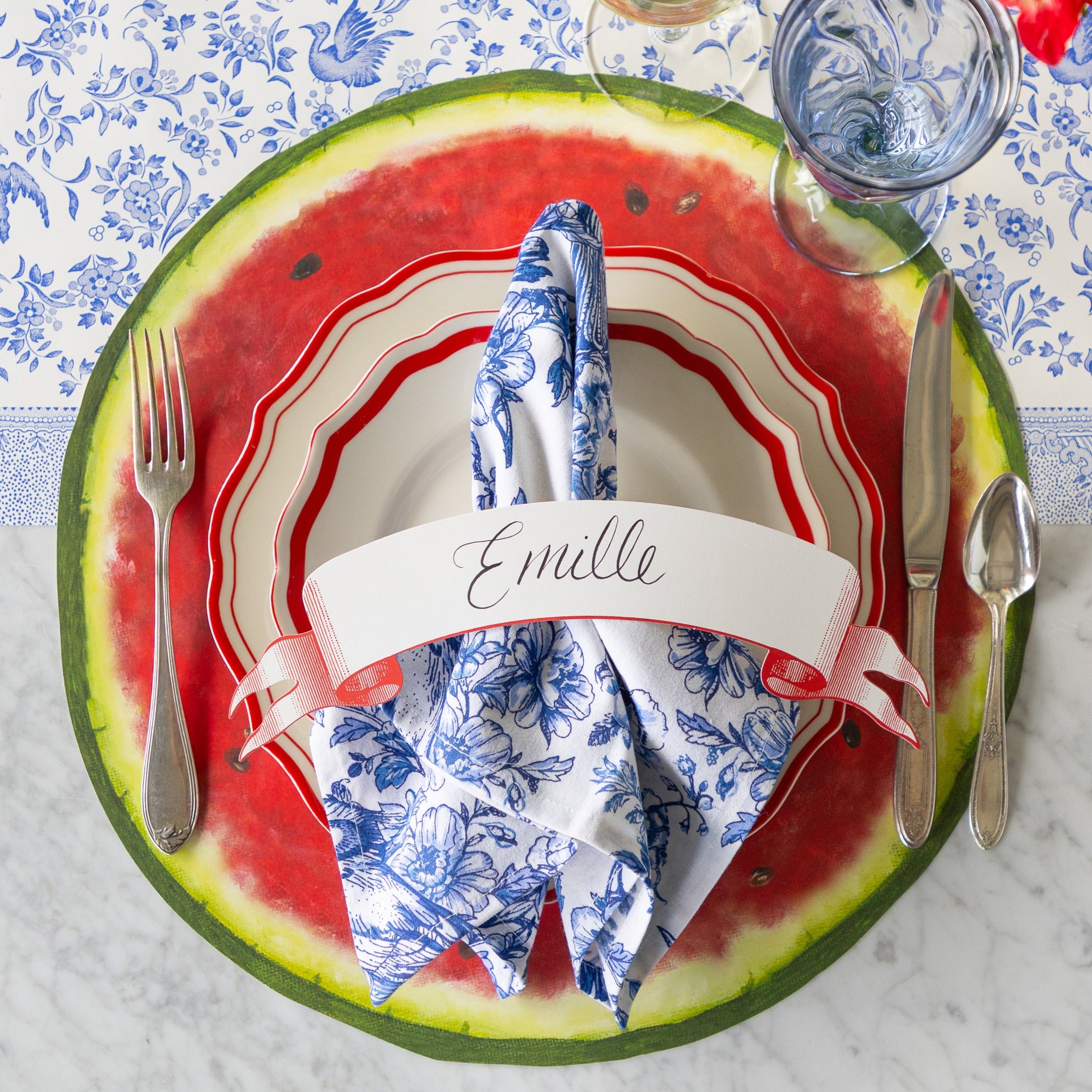 A summery place setting featuring the Die-cut Watermelon Placemat, red scalloped plates, and a blue floral napkin with a Classic Red Banner Table Accent atop on the Blue Regal Peacock Runner.