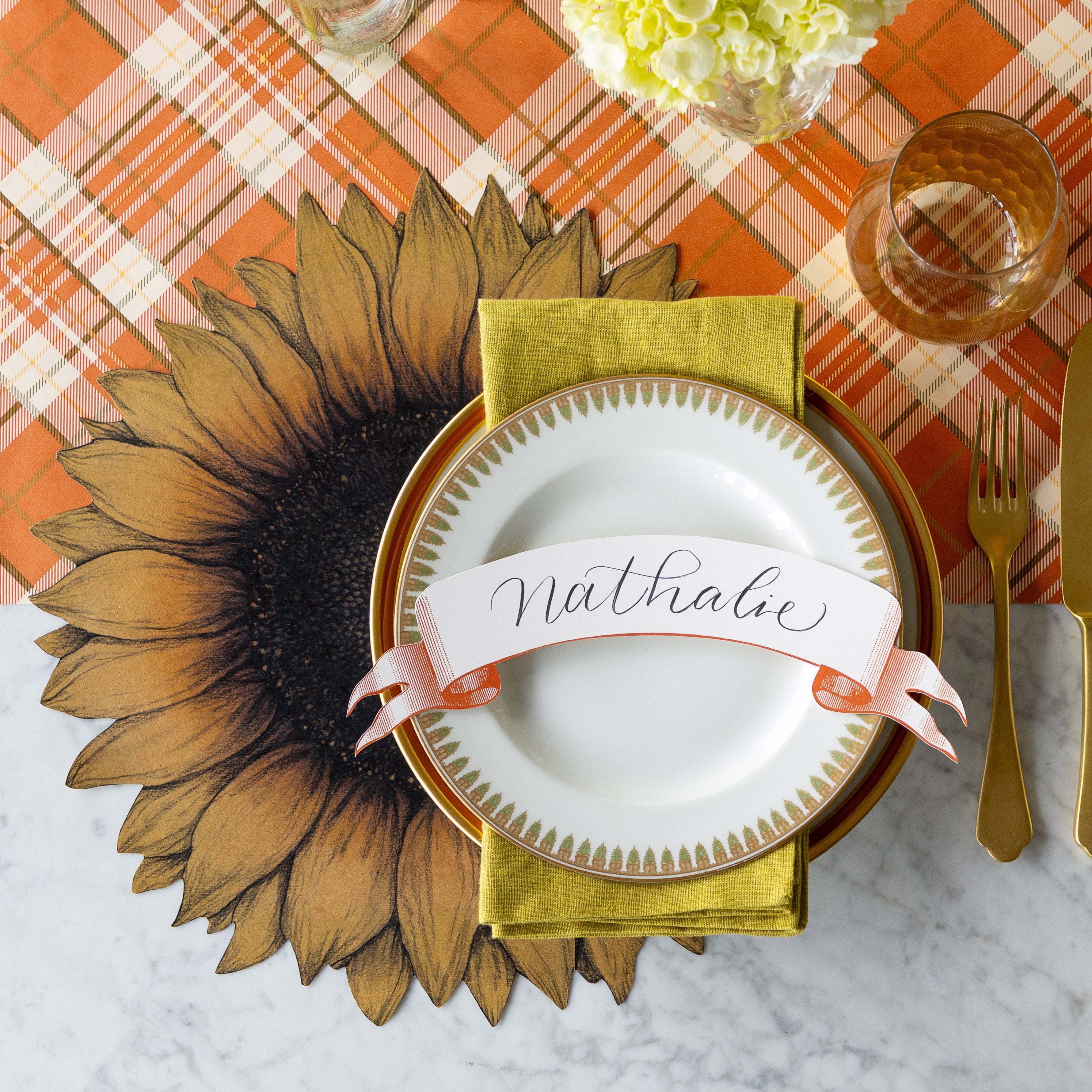 A place setting featuring the Die-cut Vintage Sunflower Placemat offset to the left under a green napkin in between vintage dinnerware with the Classic Orange Banner Table Accent reading "Nathalie" atop, and Orange Plaid Runner underneath.