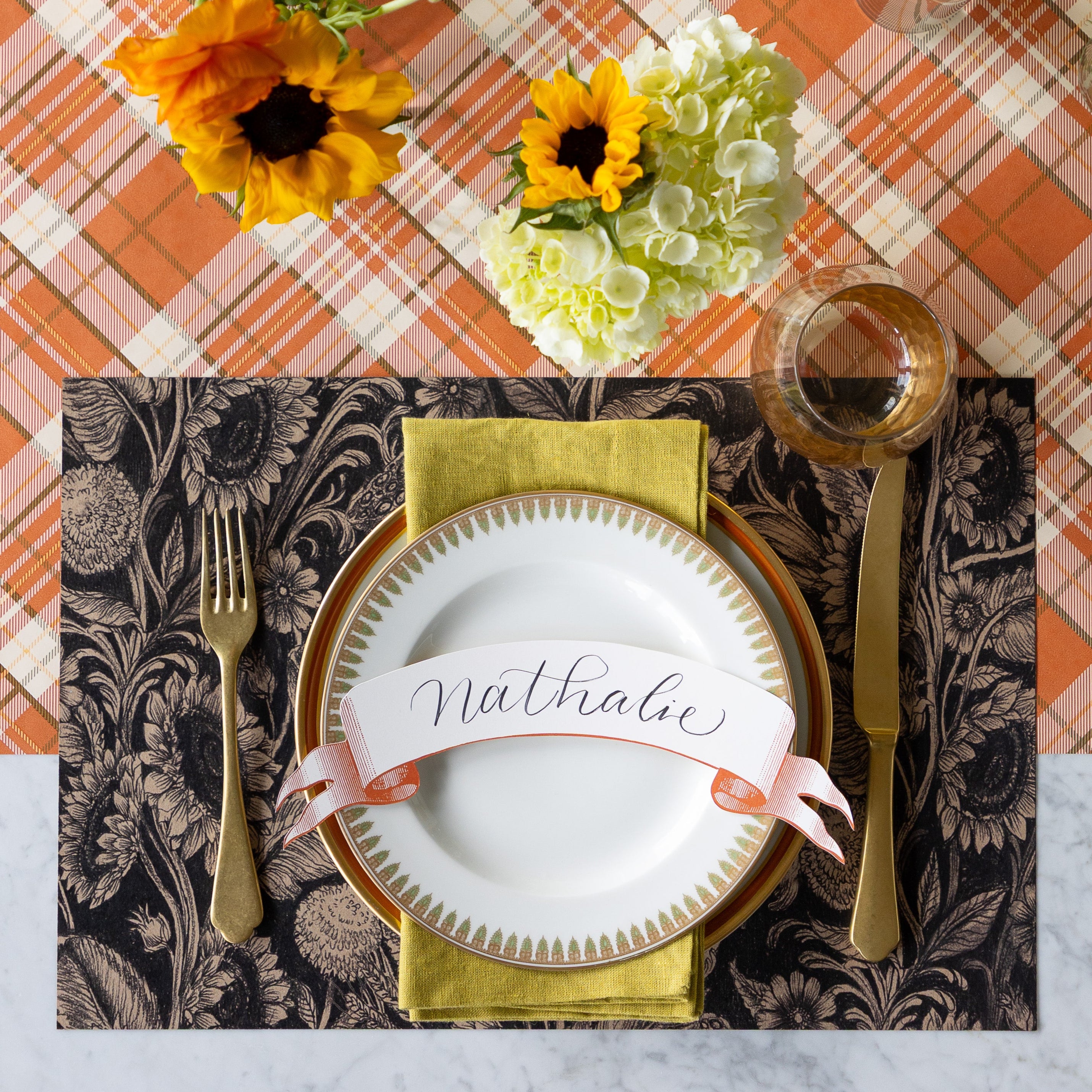 A table setting with fresh fall flowers, featuring the Sunflower Field Placemat under vintage dinnerware with the Orange Classic Banner reading "Nathalie" atop, gold flatware, green napkin and Orange Plaid Runner underneath.