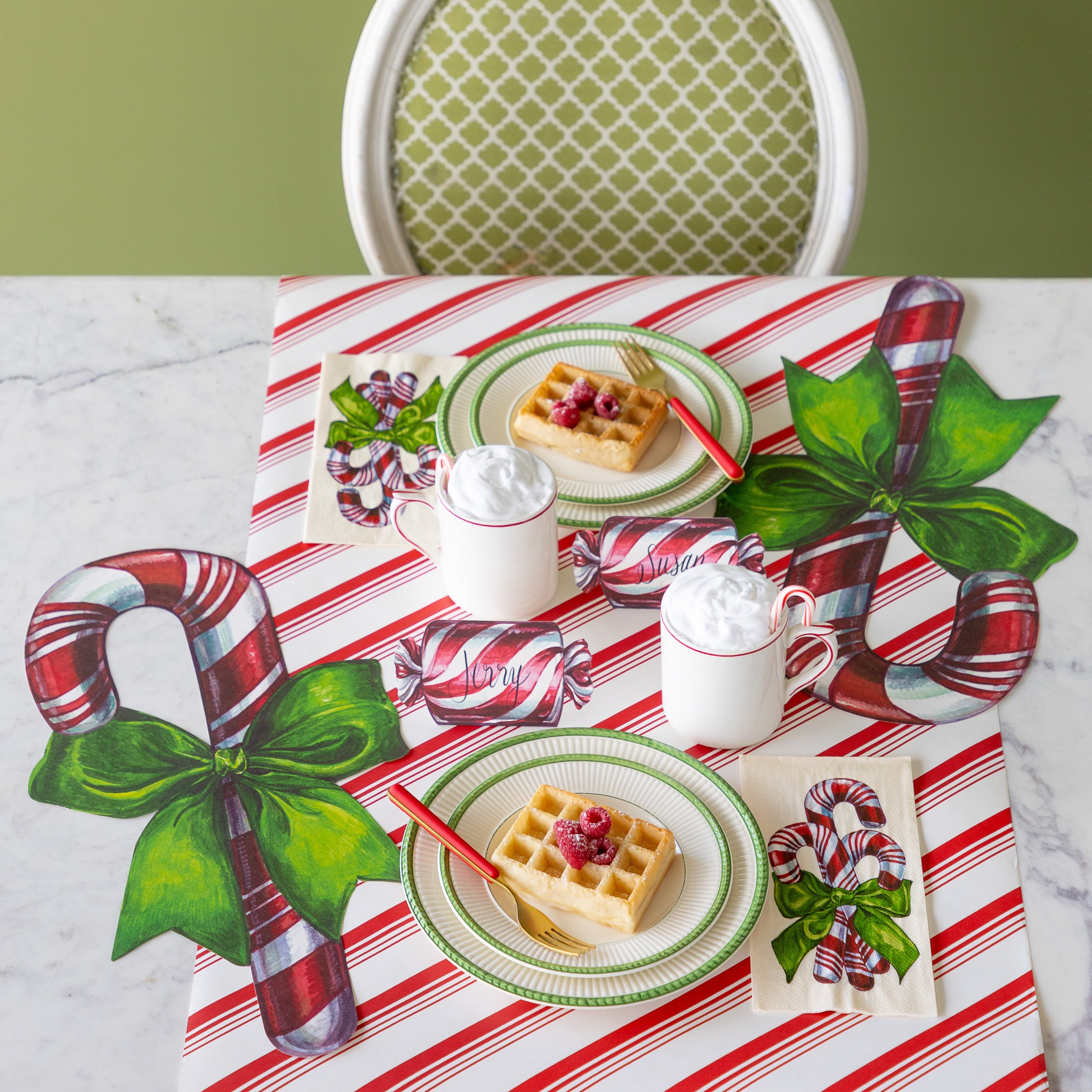 A festive table setting for two featuring the Die-cut Candy Cane Placemat next to green rimmed plates with waffles, Candy Cane Guest Napkins, cups filled with whip cream and a candy cane, and the Peppermint Stripe Runner underneath.