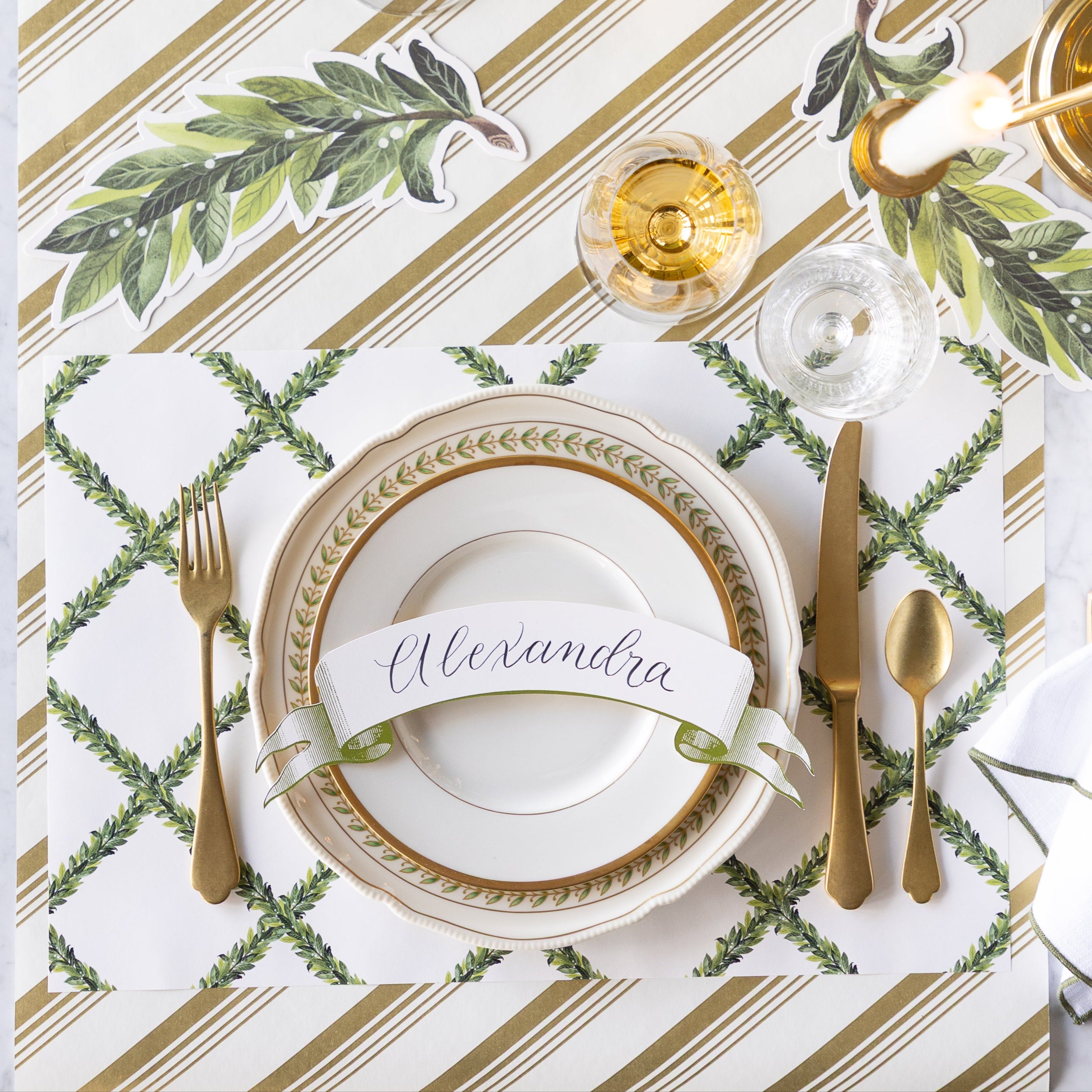 A formal place setting with gold cutlery, a wine glass, vintage plates with the Classic Moss Banner Table Accent reading "Alexandra" atop, Laurel Table Accents scattered on the table, and Laurel Wreath Placemats on the Gold Peppermint Stripe Runner.