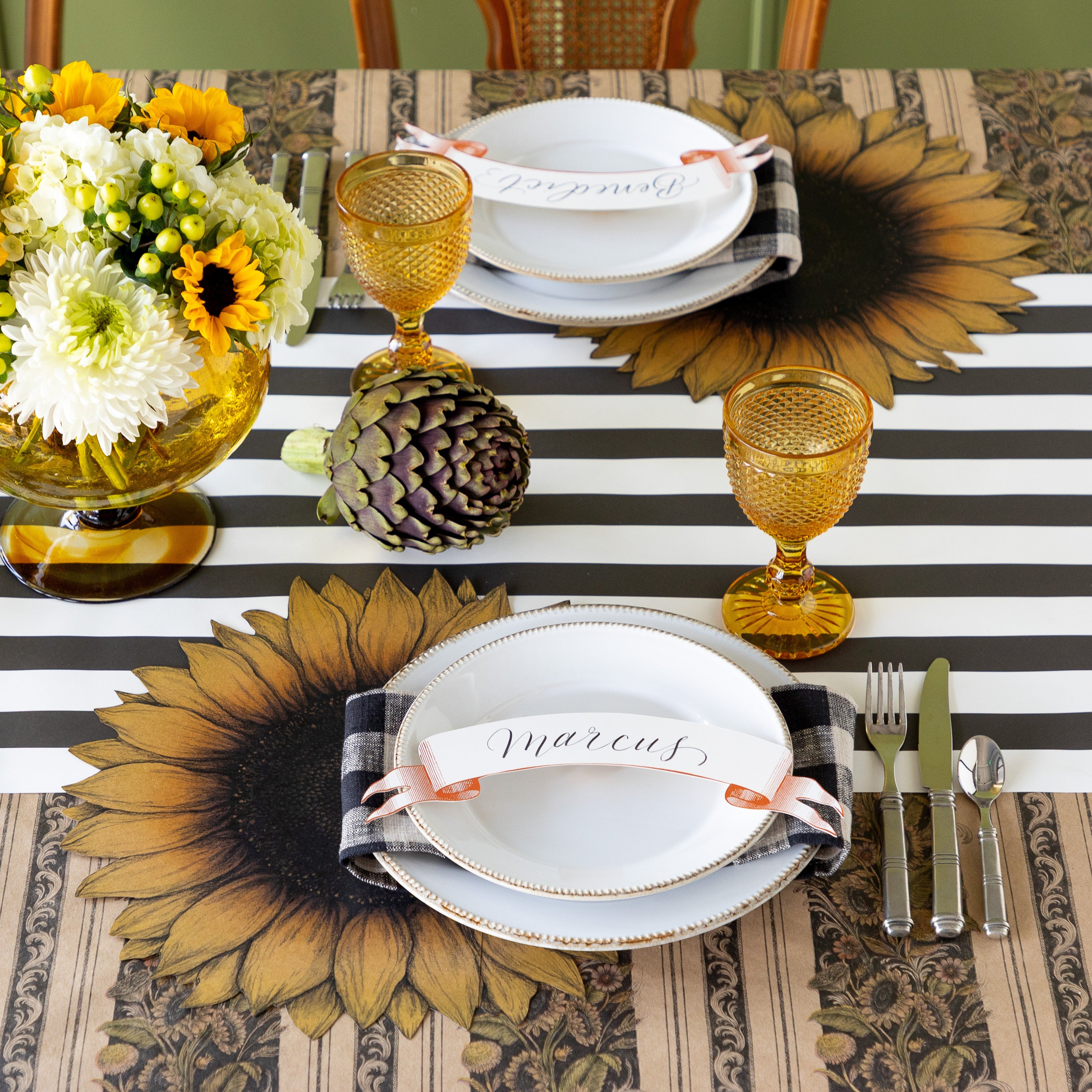 A table setting with fresh flowers centerpiece and an artichoke featuring the Die-cut Vintage Sunflower Placemat under white dinner plates with the Classic Orange Banner Table Accent atop, Vintage Black Sunflower Stripe Runner rolled vertically and Black Classic Stripe Runner rolled out horizontally underneath.