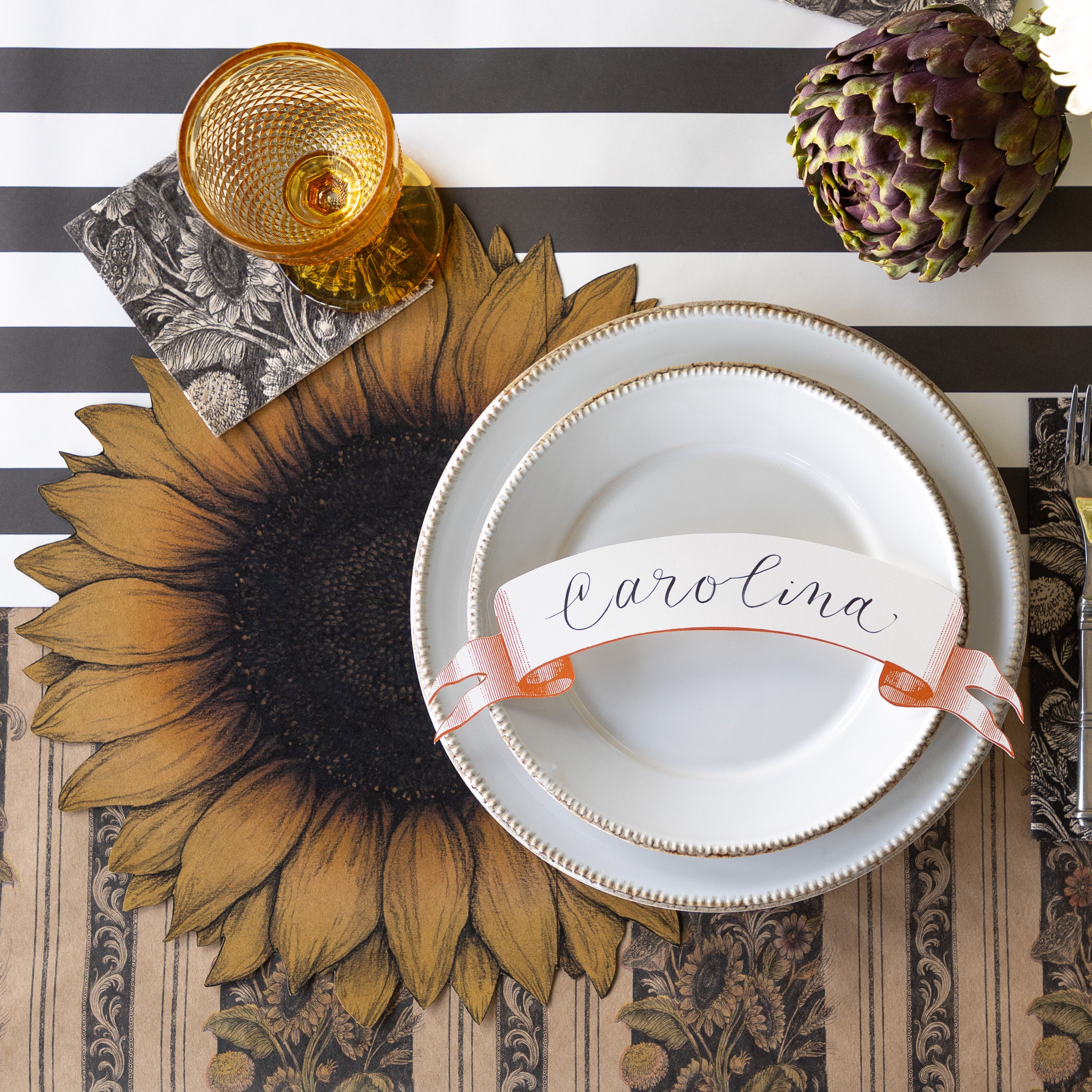 A table setting with an artichoke, featuring the Die-cut Vintage Sunflower Placemat, Vintage Sunflower Napkins, two white plates with the Classic Orange Banner Table Accent reading "Carolina" atop, Vintage Black Sunflower Runner rolled out vertically and Black Classic Stripe Runner horizontally underneath.