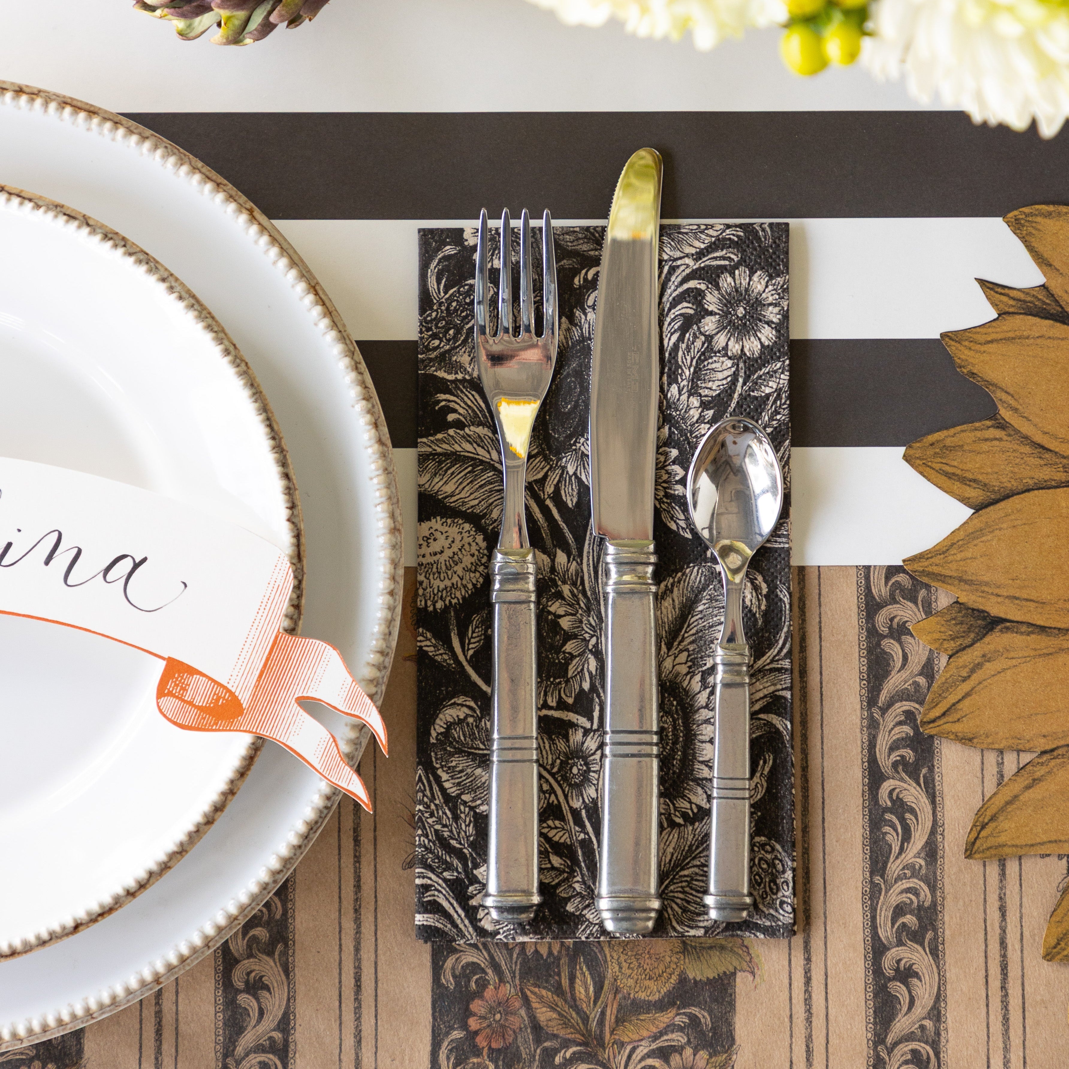 A place setting with the Vintage Sunflower Guest Napkin under silverware next to it, on top of the Vintage Black Sunflower Runner and Black Classic Stripe Runner.