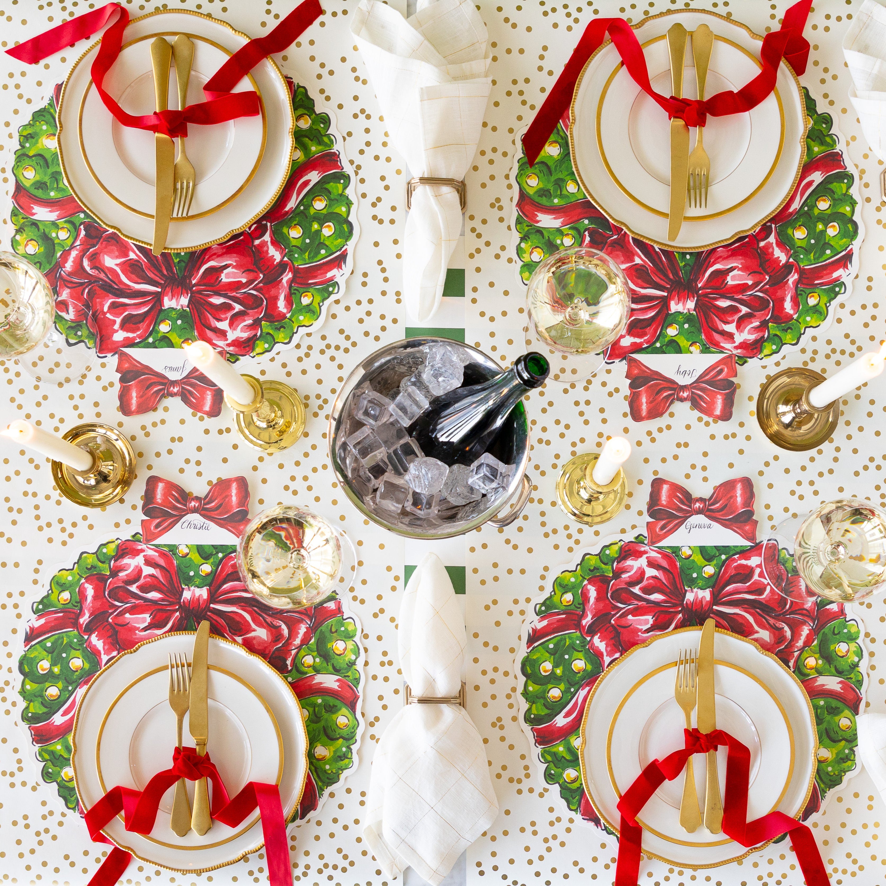 A table setting for four with lit candles and an ice bucket of champagne, featuring the Die-cut Holiday Wreath Placemats, gold rimmed dinnerware, gold flatware tied in a red velvet ribbon, and napkins in the Napkin Ring with Place Card Holder on the Gold Confetti and Dark Green Classic Stripe Runner.