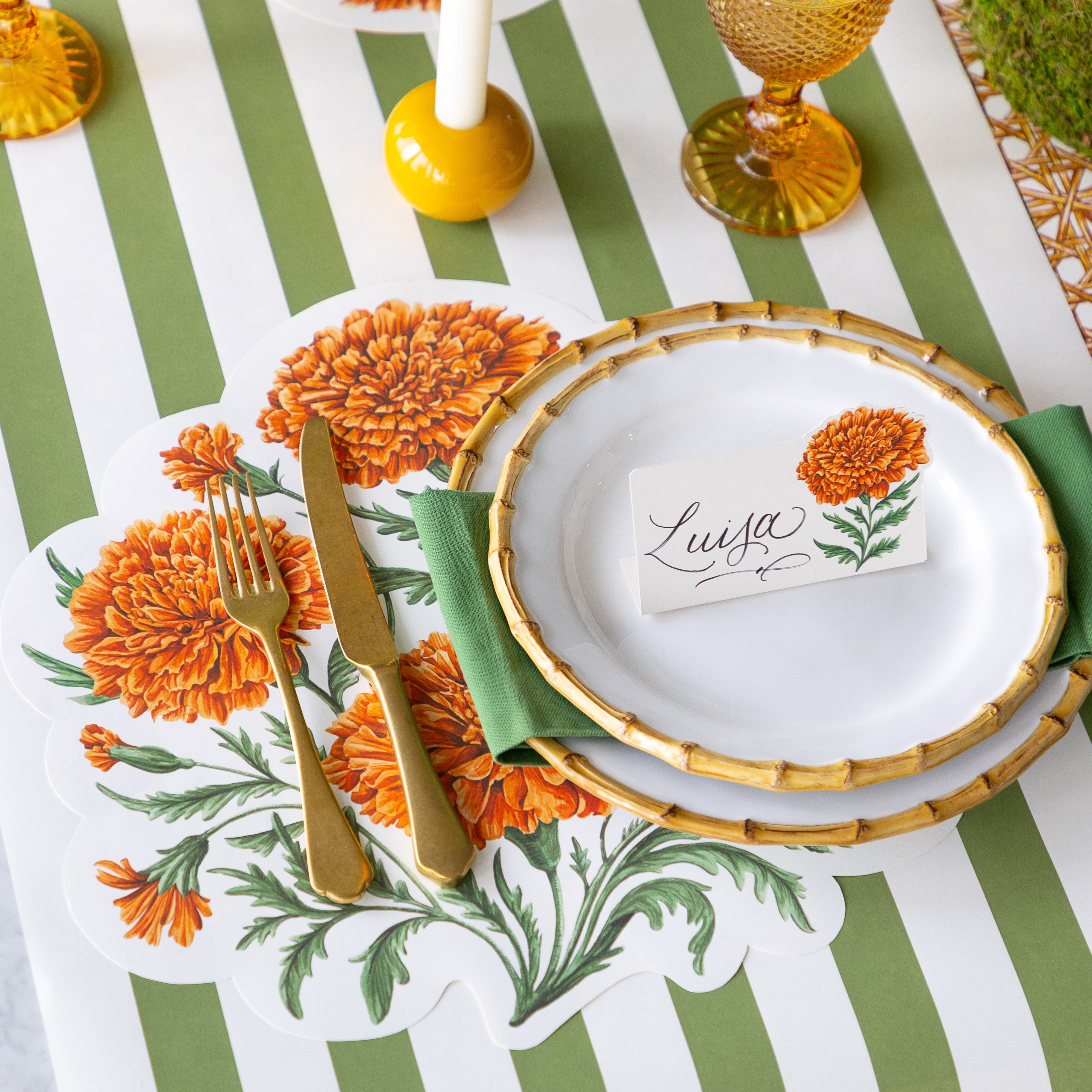 An elegant place setting featuring the Die-cut Marigold Bouquet Placemat offset to the left under the Mepra Gold fork and knife, a green napkin in between Juliska Bamboo Dinner Plates with the Marigold Bloom Place Card on top, and Classic Moss Stripe Runner underneath.