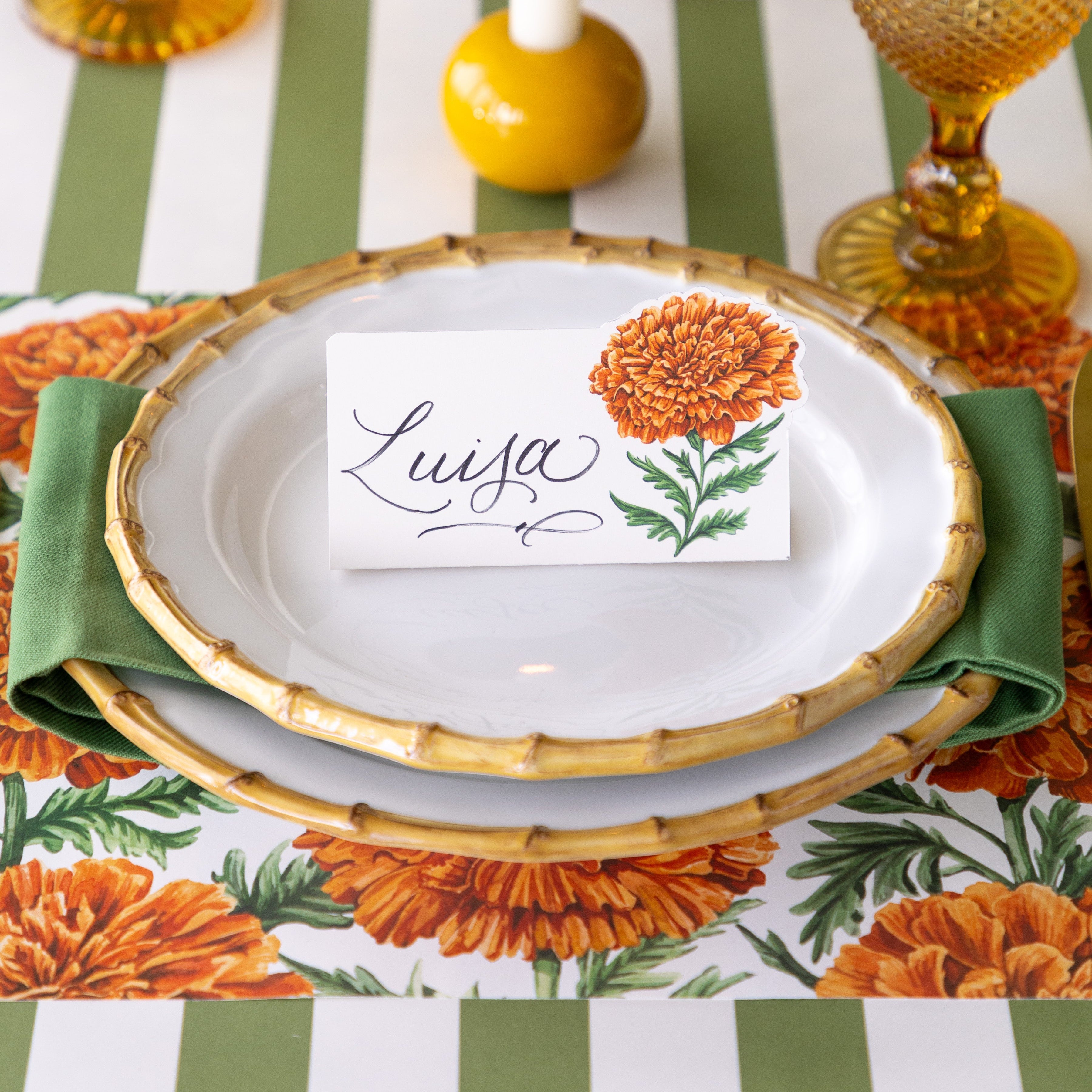 A place setting featuring the Marigold Bloom Place Card reading "Luisa" atop bamboo plates with the Marigold Harvest Placemat and Moss Classic Stripe Runner underneath.