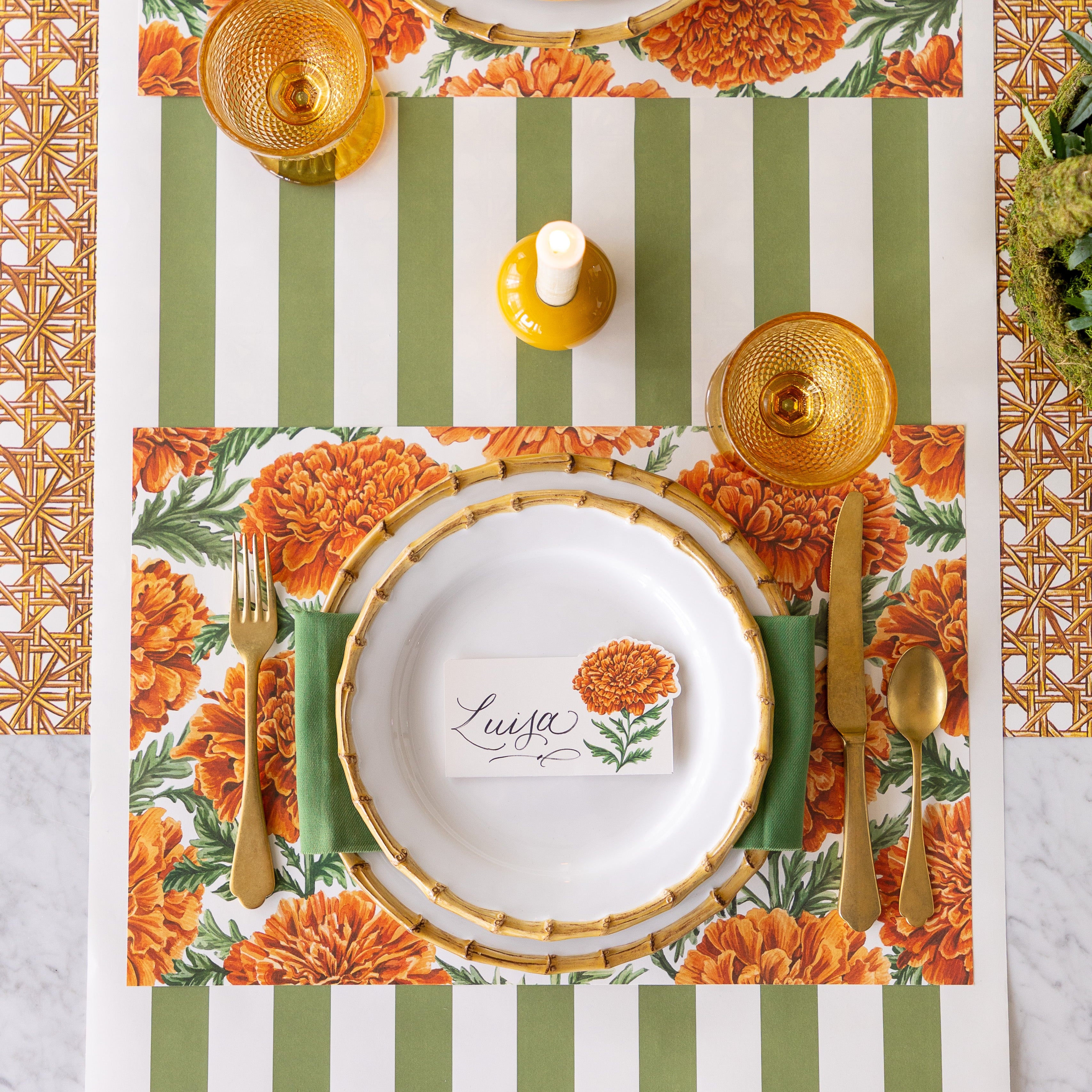 A set table with a lit candle featuring the Marigold Harvest Placemat under bamboo dinnerware with the Marigold Bloom Place Card reading "Luisa" atop, gold flatware, an orange glass, and Moss Classic Stripe Runner vertically on the table and Rattan Reave Runner horizontally underneath.
