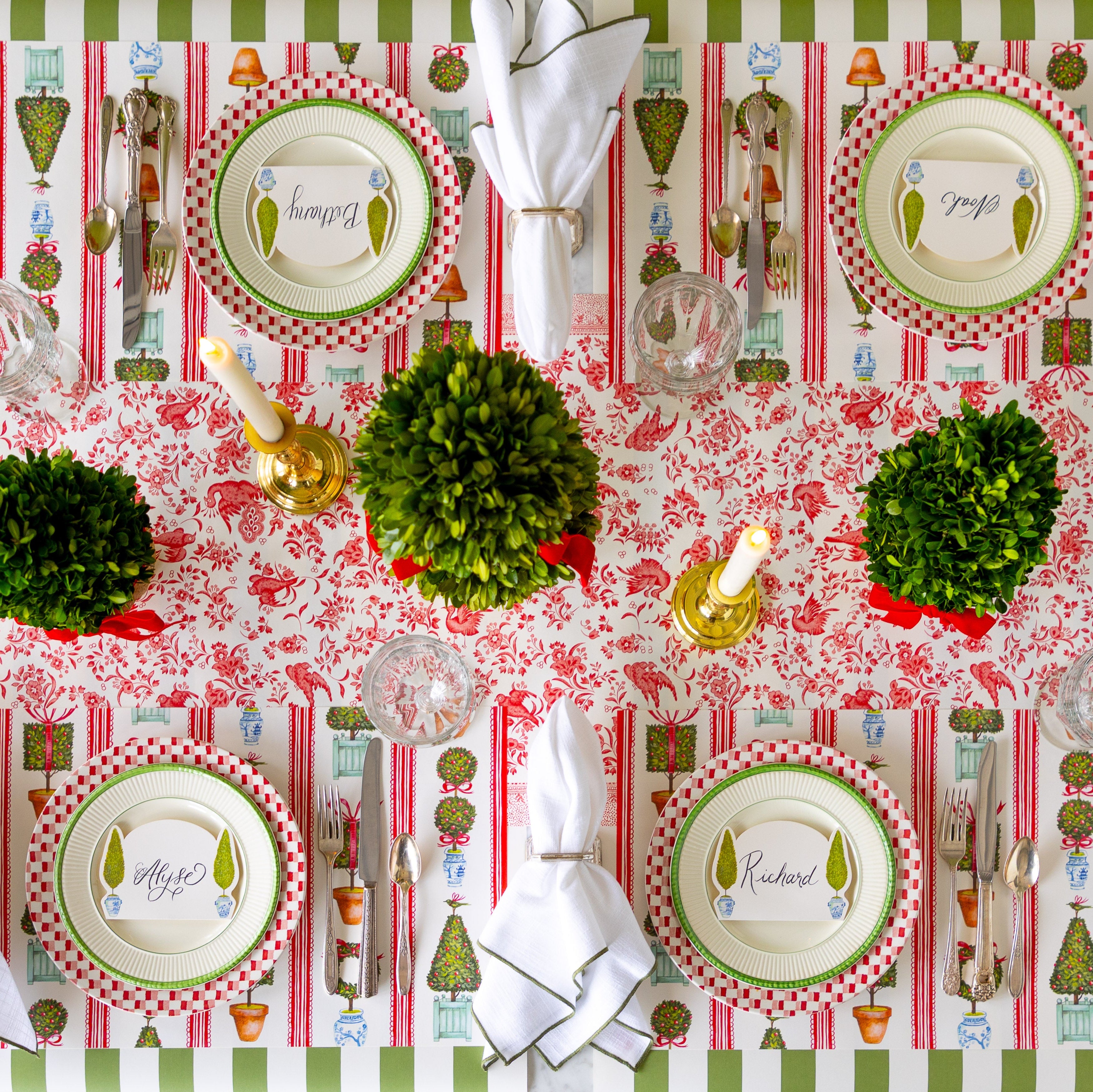 A table set for four with boxwood topiary and candle center pieces, featuring the Red Regal Peacock Runner under Holiday Topiary Placemats with red-and-white checkered and green rimmed plates, vintage silverware and Topiary Place Cards atop.