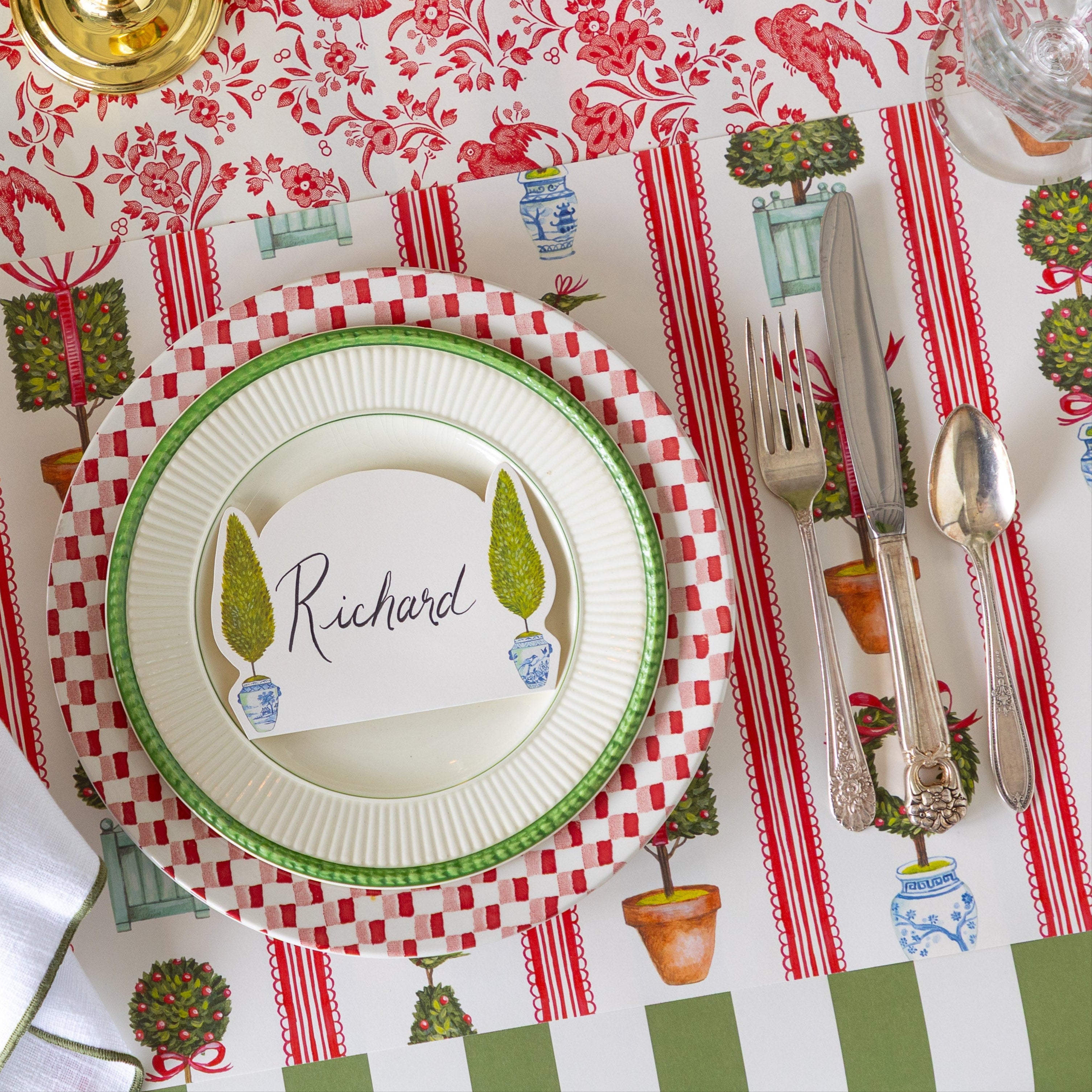 An elegant place setting featuring the Holiday Topiary Placemat, dinner plates with the Topiary Place Card on top, Moss Classic Stripe Runner running vertically and Red Regal Peacock Runner running horizontally underneath.