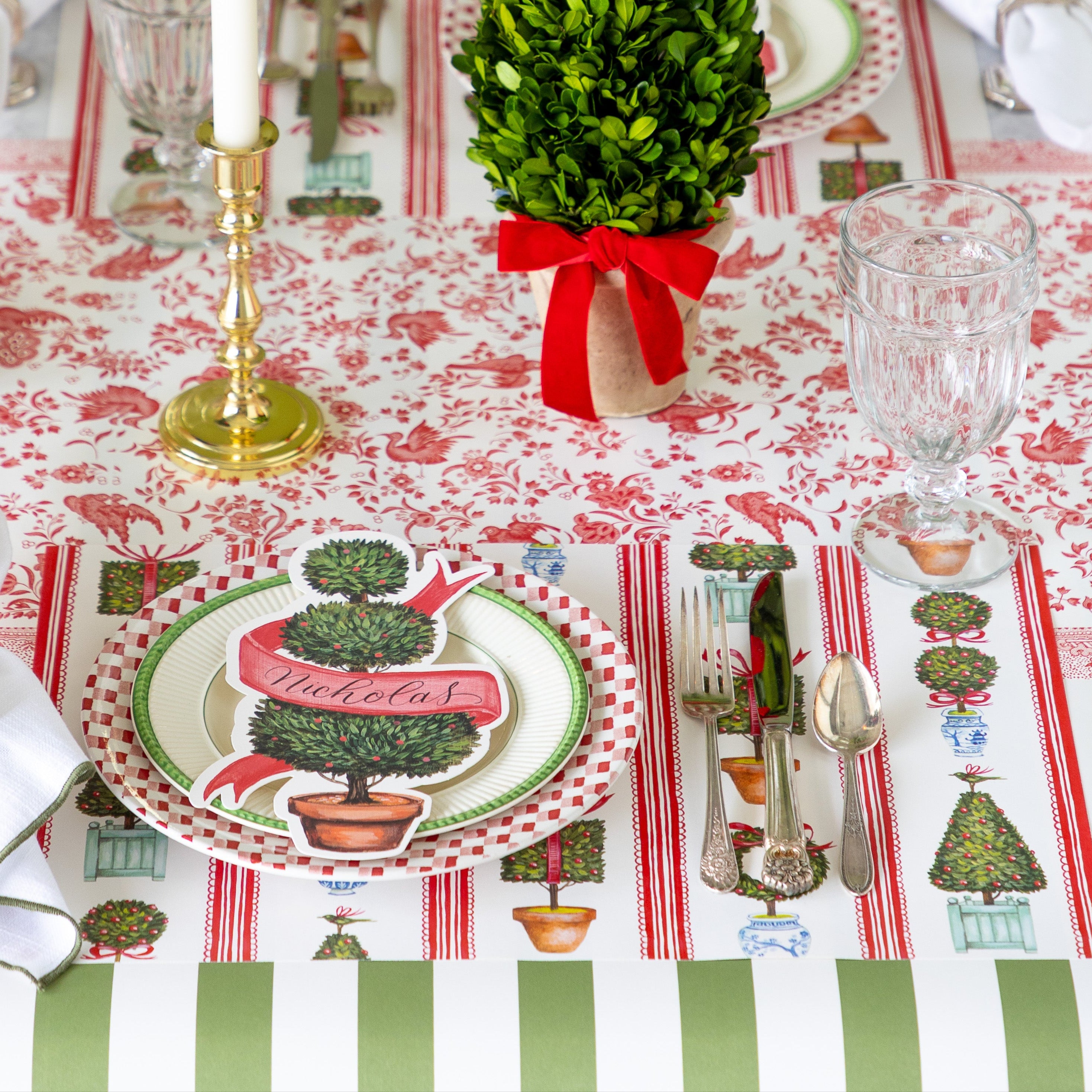An elegant place setting featuring the Holiday Topiary Placemat, dinner plates with the Holiday Topiary Table Accent on top, Moss Classic Stripe Runner running vertically and Red Regal Peacock Runner running horizontally underneath.